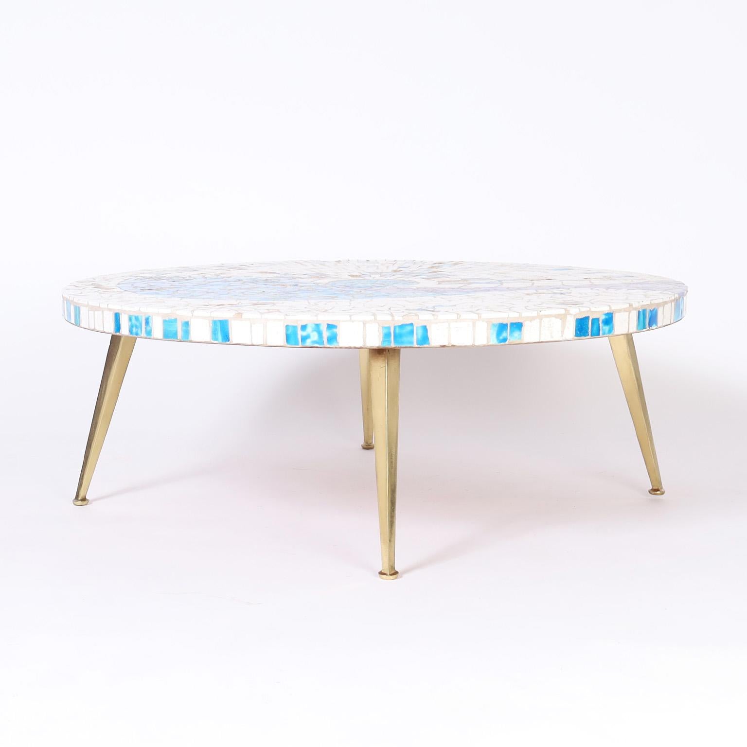 Striking mid century round coffee table with a mosaic tile top having an alluring stylized aquatic motif on elegant tapered brass legs.