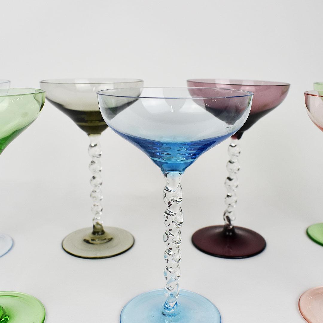 Collection of 7 multi-color pastel coupe glass drinking glasses with twisted stems. The body of the glasses are short with a wide mouth in the shape of a typical coupe glass, each in a different color. The stems are in a transparent glass, with a