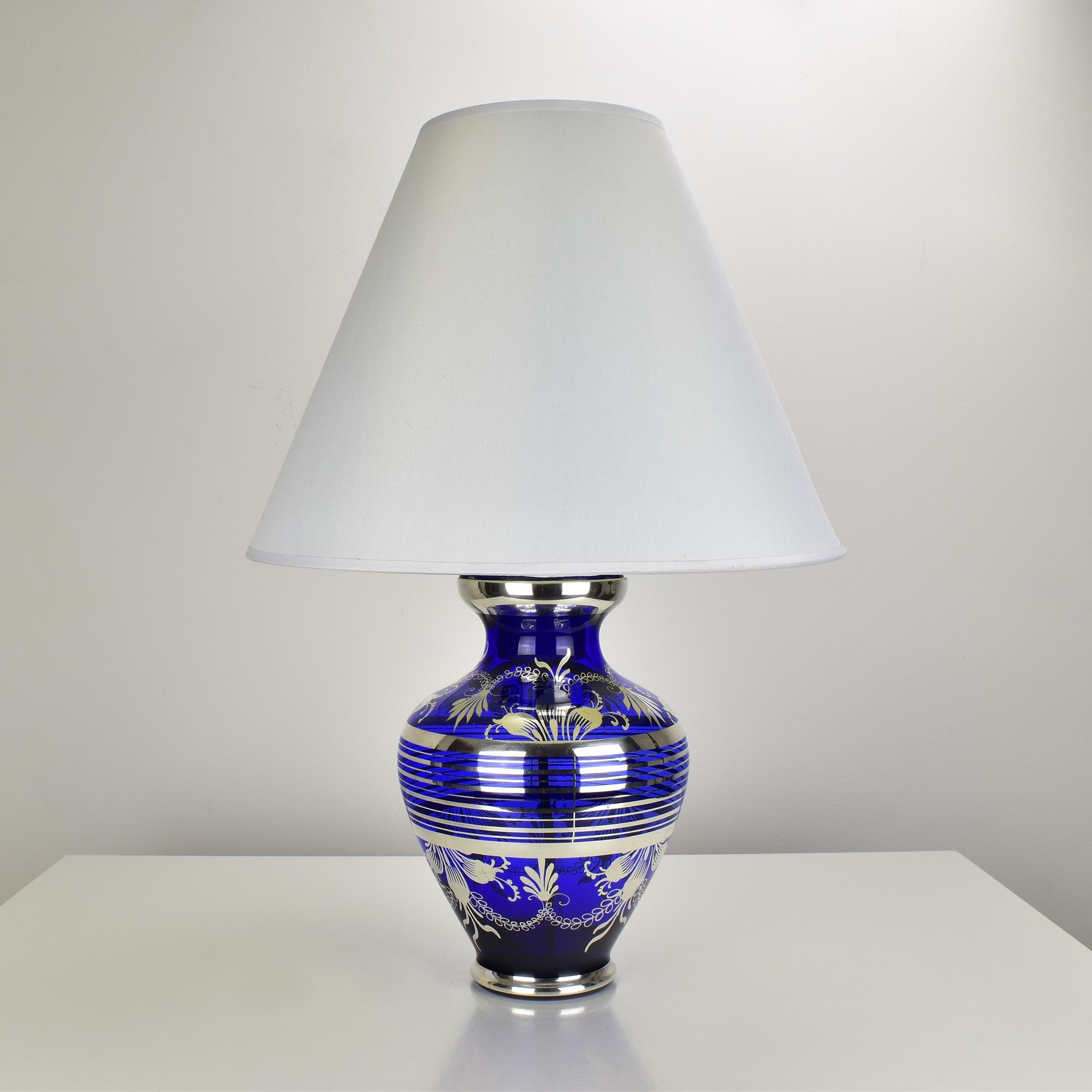 This striking Murano glass table lamp from the 1960s features a vivid cobalt blue body with an elegant silver overlay decoration, attributed to Balboa. The intricate silver pattern adds a luxurious and timeless appeal to the piece, making it a