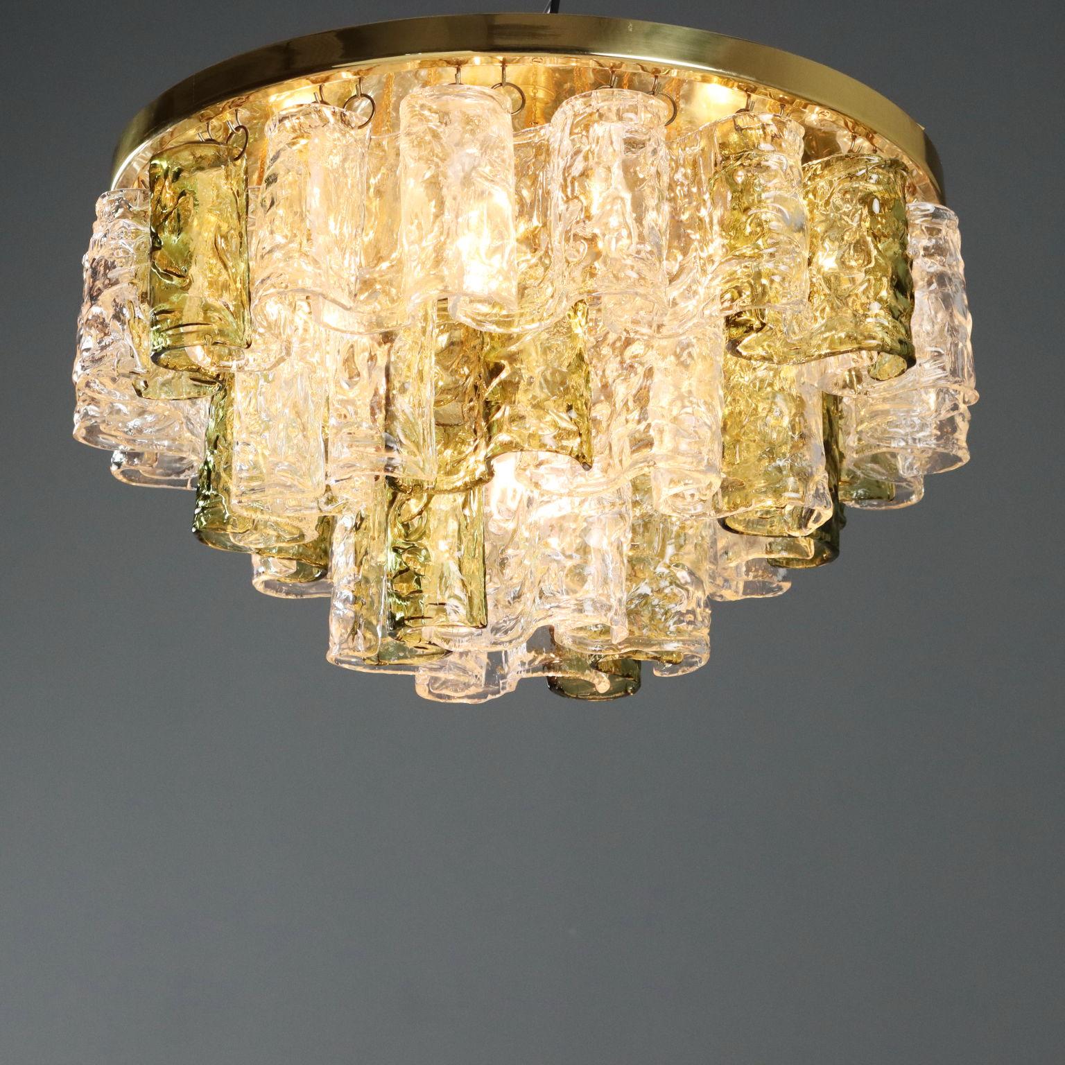 Questo lampadario esemplifica la caratteristica lavorazione artigianale del vetro di Murano. Il design è caratterizzato da elementi cilindrici in vetro trasparente e ambrato sospesi da una struttura in ottone, che creano un effetto di luce