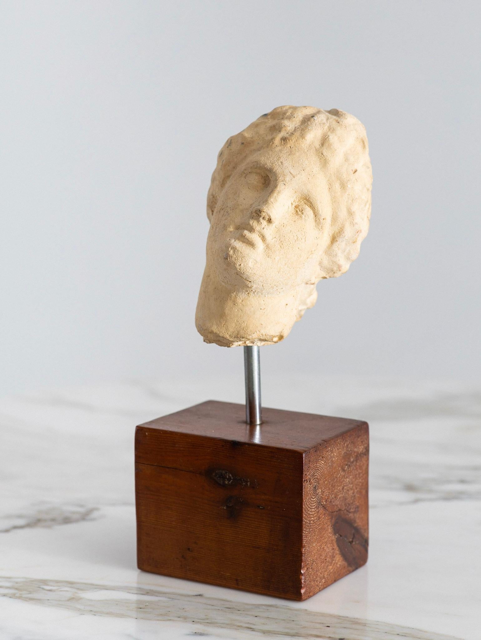 A mid century Objet d’art. A museum style replica of an ancient Greek or Roman statue fragment. Plaster female head and neck form mounted on a wood plinth base. Unmarked.