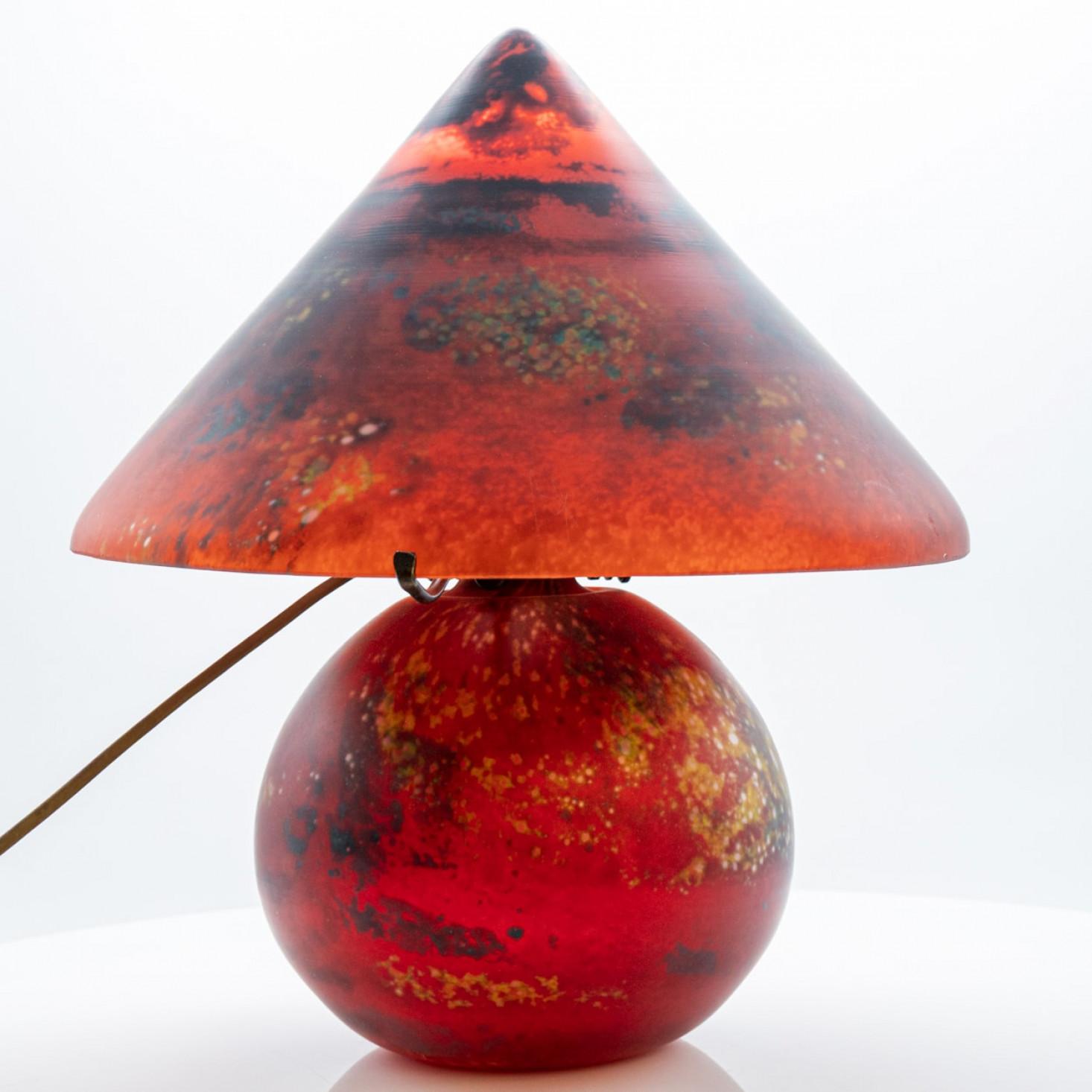 Una lámpara de mesa única y artística con forma de seta. La lámpara tiene una base redonda de cristal rojo con un patrón moteado de acentos naranjas, amarillos y oscuros, que le da un aspecto ardiente, casi planetario. La pantalla es cónica y de