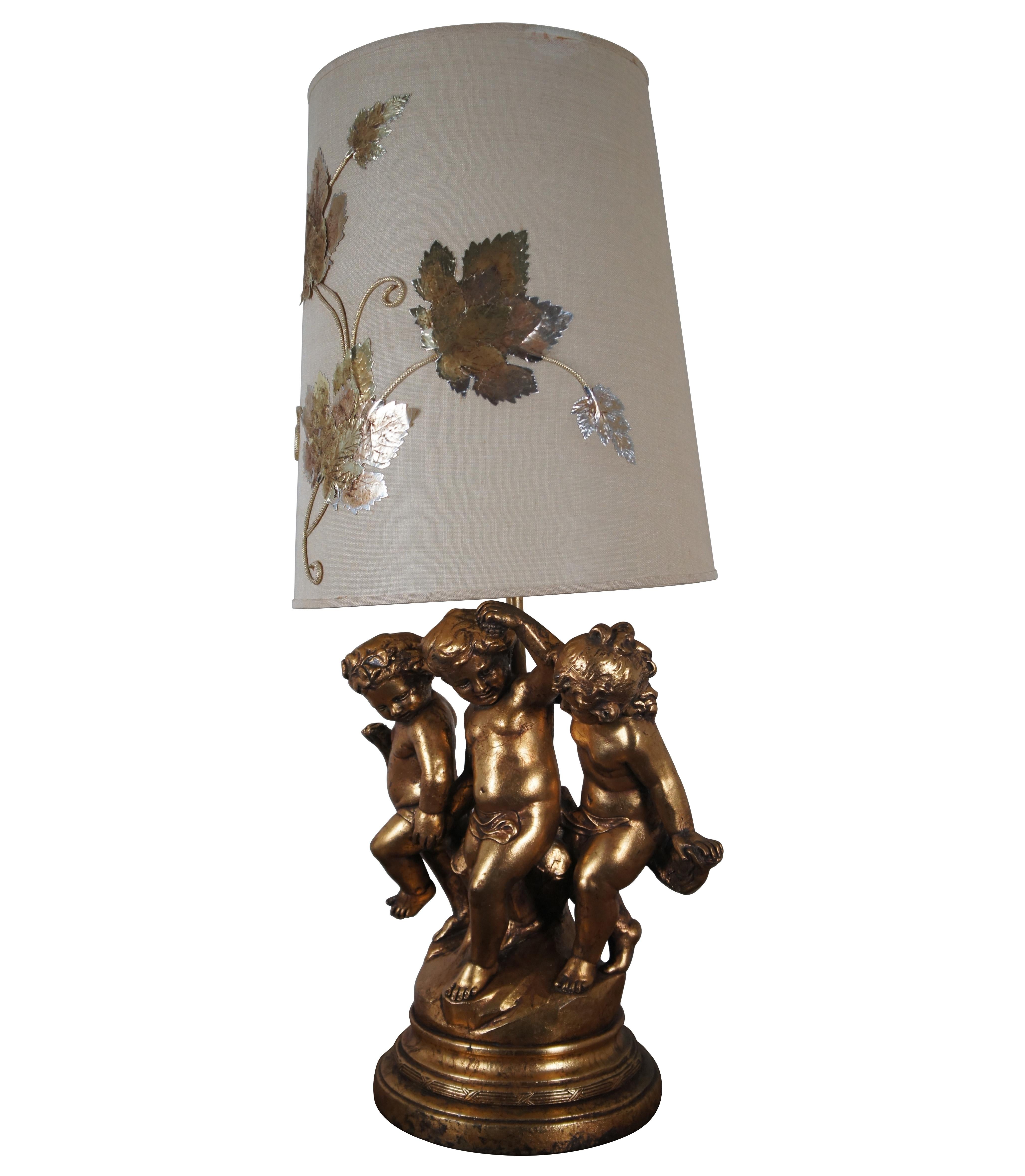 A large and impressive / monumental Mid-20th Century gilded toleware sculptural table lamp showing three Neoclassical putti figures / wingless cherubs on a round plinth. Marked Original Art on back side. Paired with immense linen shade accented with