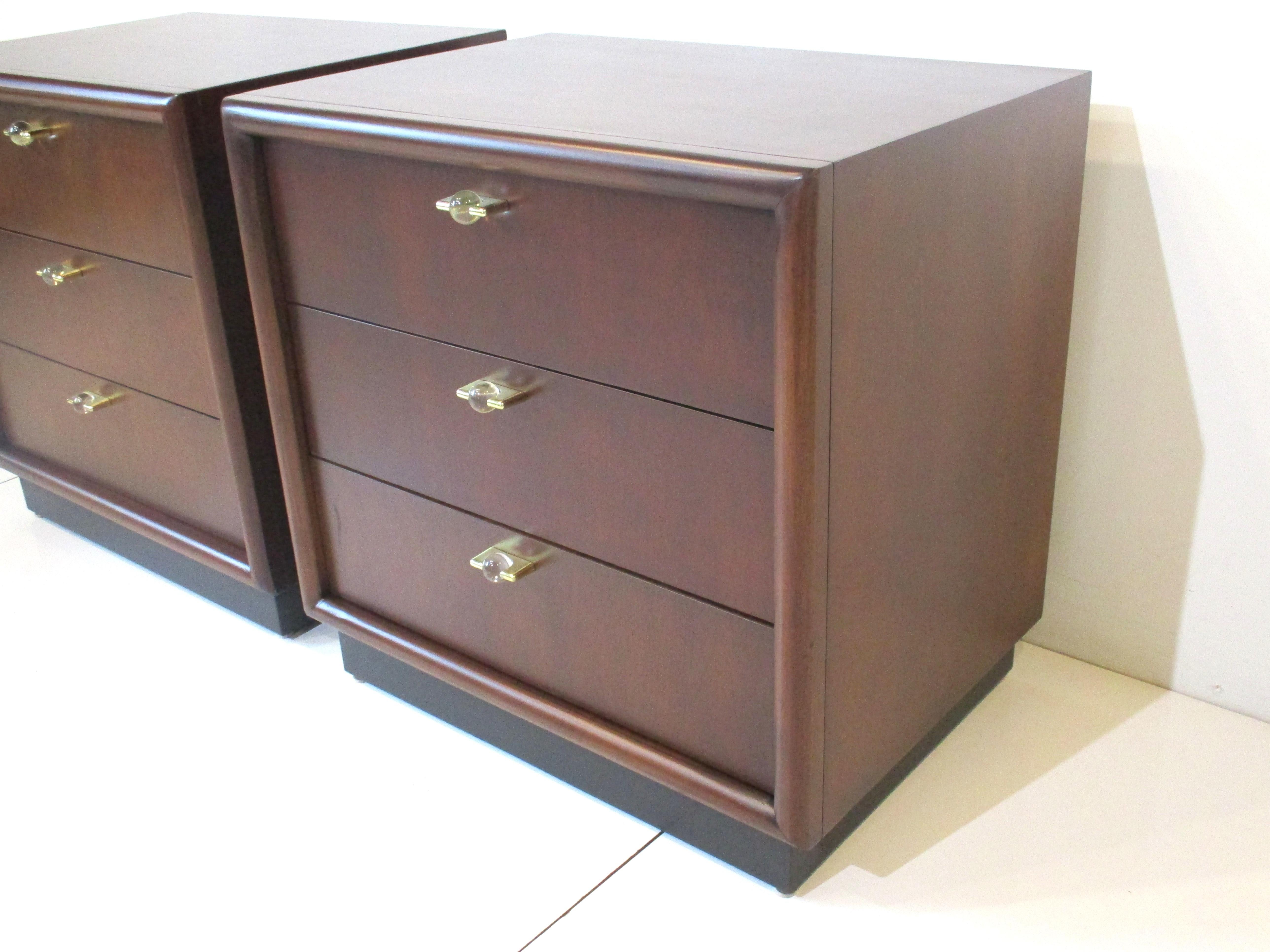 A pair of very well crafted nightstands in medium dark toned mahogany, three drawers with flat brass pulls with clear round Lucite balls to the front. The lower area has a satin black kick styled base and retains the manufactures label by the