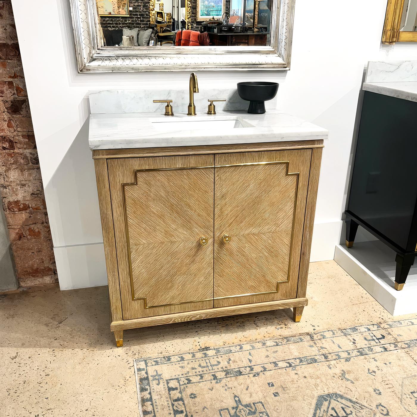 La finition cérusée claire met en valeur le grain naturel du bois, tandis que les détails géométriques des panneaux et les accents en laiton ajoutent une sophistication architecturale. Un luxueux plateau en marbre blanc de Carrare couronne la pièce,