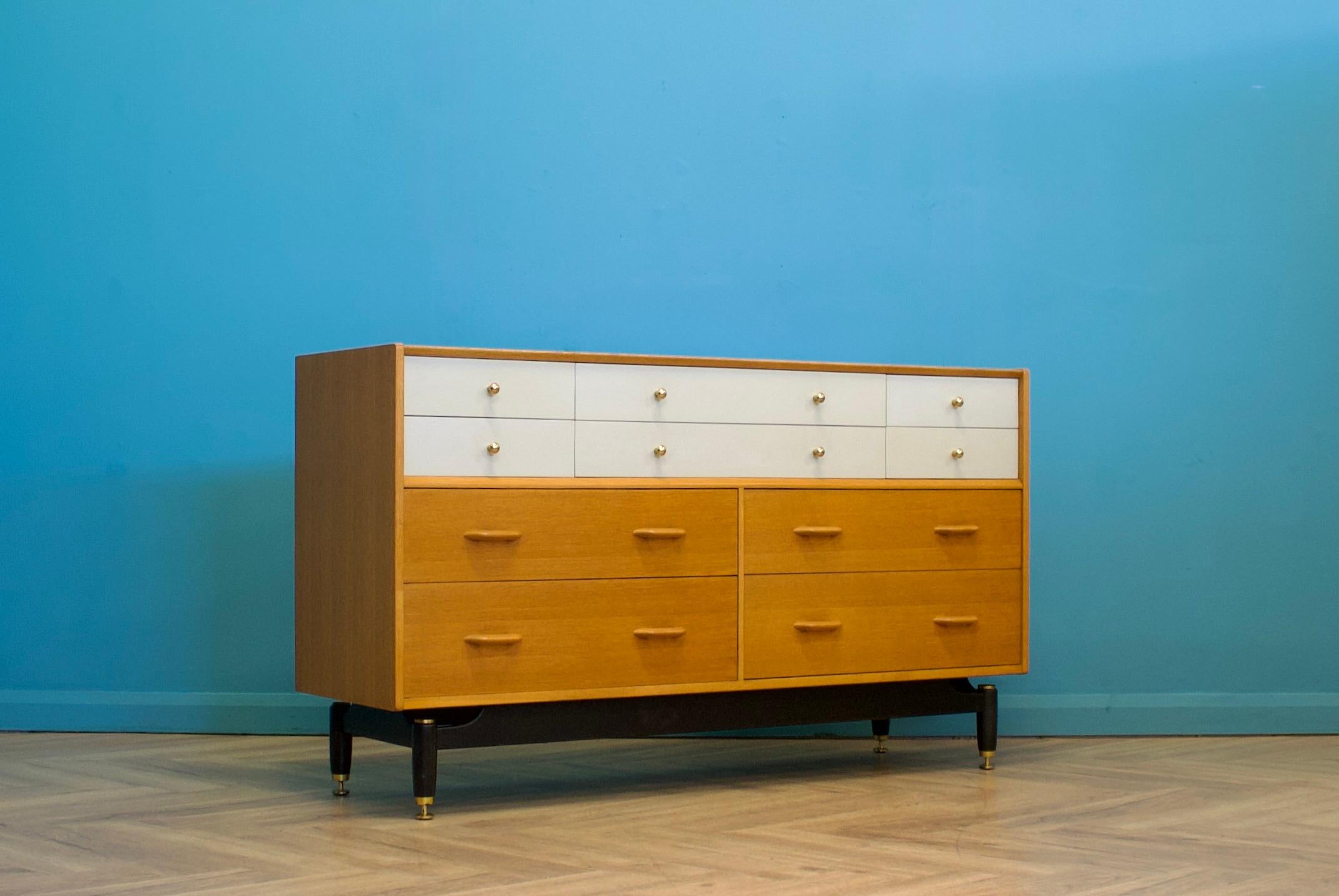 This large oak chest of drawers from G Plan (E Gomme) in the China White range dates to the 1950s. The top drawers retain their original white lacquered finish and brass handles, while the bottom set a golden oak veneer with solid oak