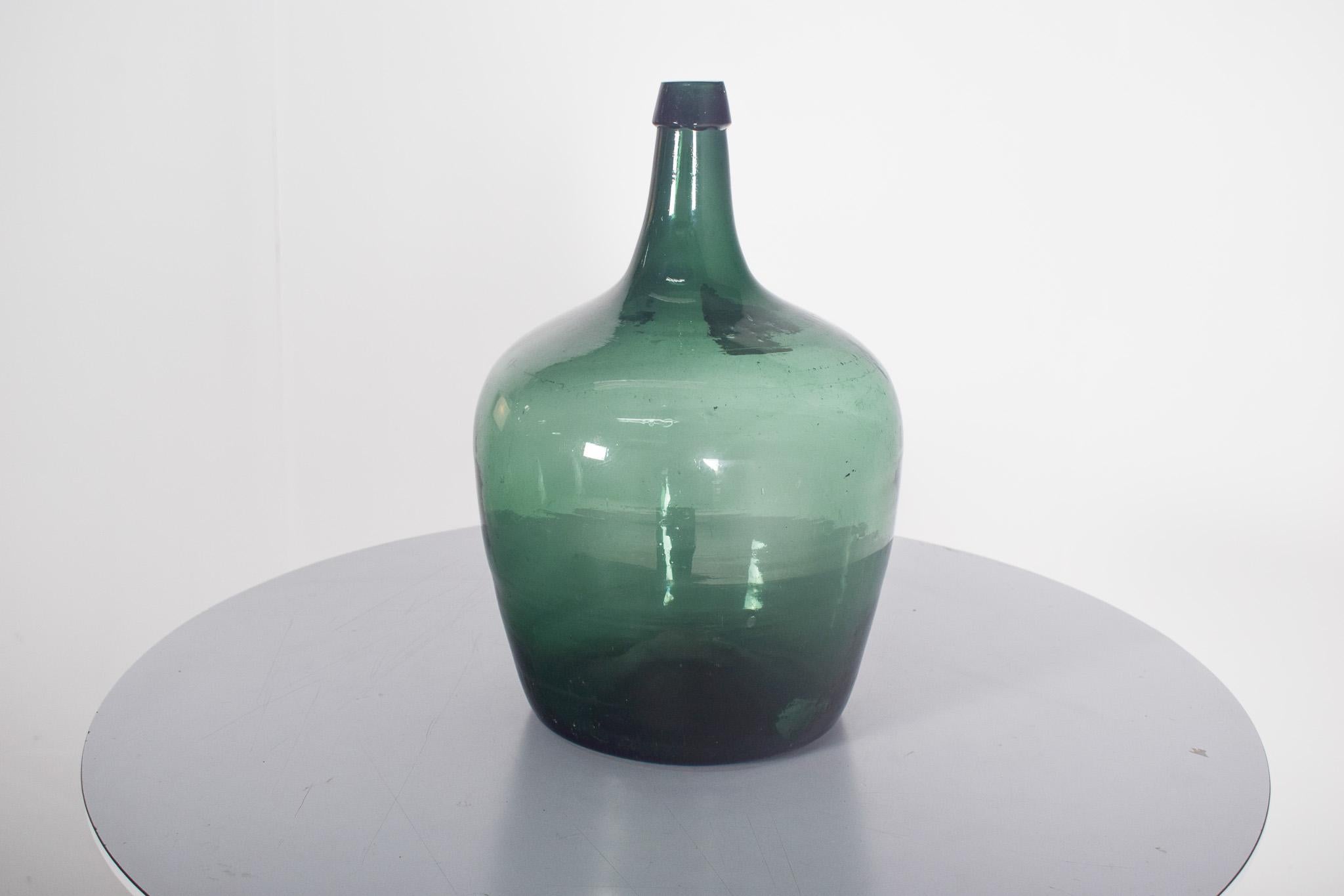 Alte grüne Glasflasche. Mit einer gewölbten Form und einer breiten Basis, die sich sanft zu einem schmalen Hals verjüngt. Das dicke und leicht unregelmäßige Glas deutet auf einen handwerklichen Herstellungsprozess hin, möglicherweise durch die