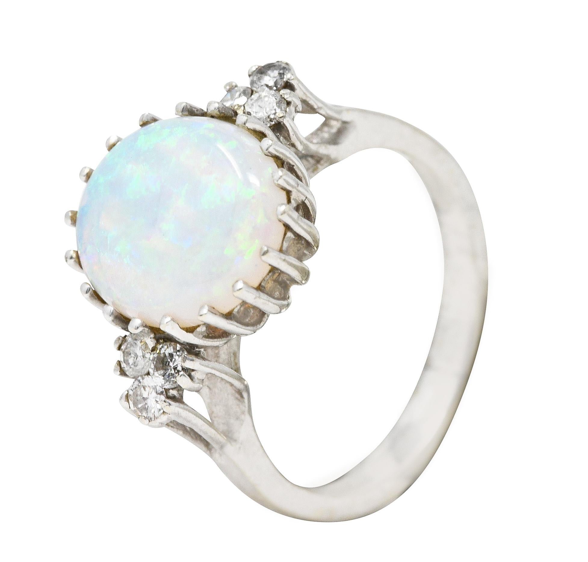 Centering an oval opal cabochon measuring 9.0 x 11.0 mm. Translucent white in body color with spectral play-of-color. Prong set in wire basket and flanked by cathedral shoulders. With clustered round brilliant cut diamonds. Prong set and weighing