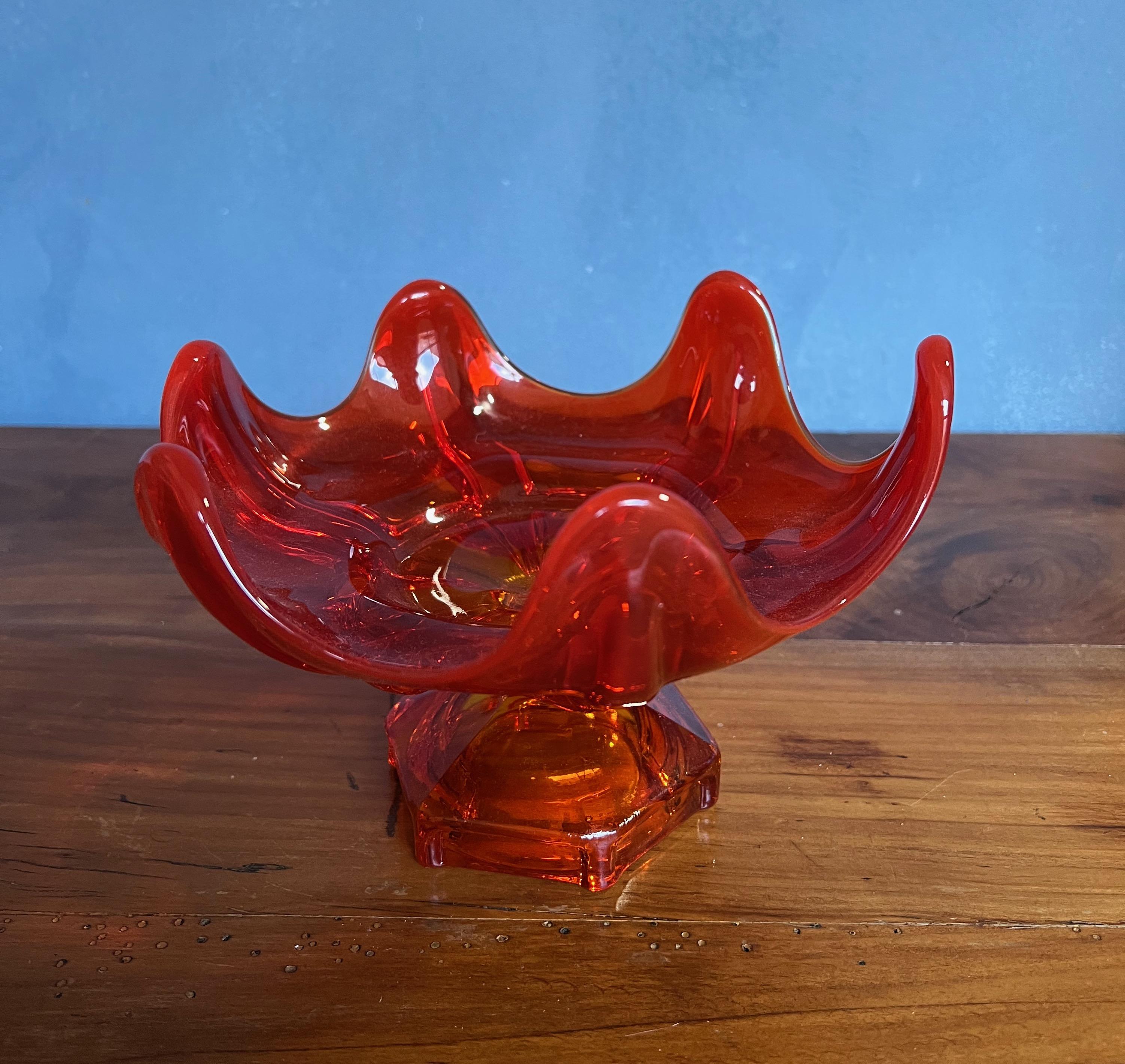 This American glass dish from the 1960s is a beautiful blend of design and craftsmanship. With a scalloped wave design and resting on a footed hexagonal base, it's a striking piece that catches the eye. The ruby/amber color of the glass adds a