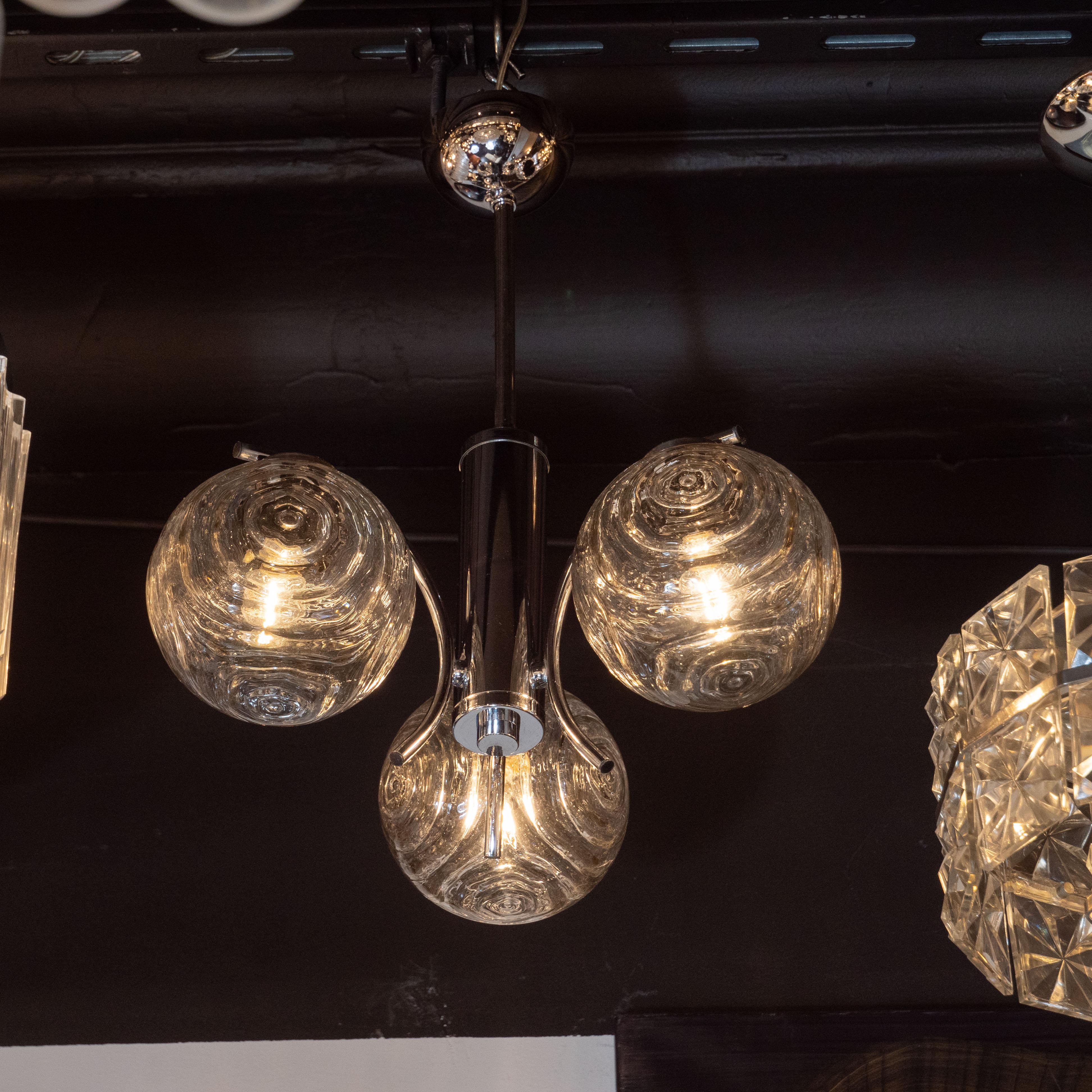 This elegant and understated Mid-Century Modern chandelier was realized in Germany, circa 1970. It features three subtly smoked translucent orbs with a concentric curvilinear pattern resembling ripples on the surface of water. Three curved arms