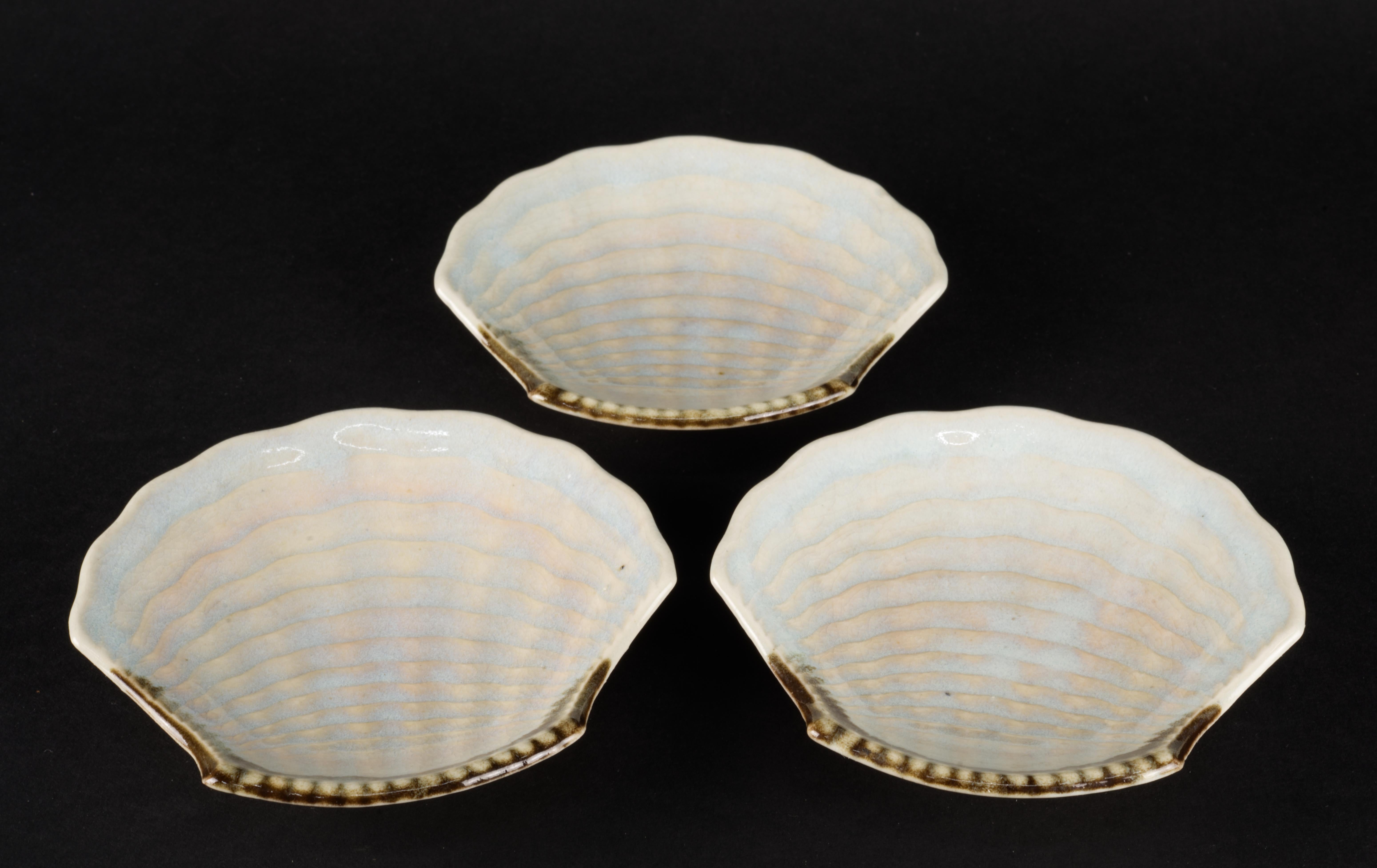 Ensemble de 3 petites assiettes ou bols en forme de coquillage, décorés à la main en glaçure craquelée dans des tons bleus et beiges avec un accent asymétrique de couleur marron terreux. Les surfaces texturées des bols sont mises en valeur par