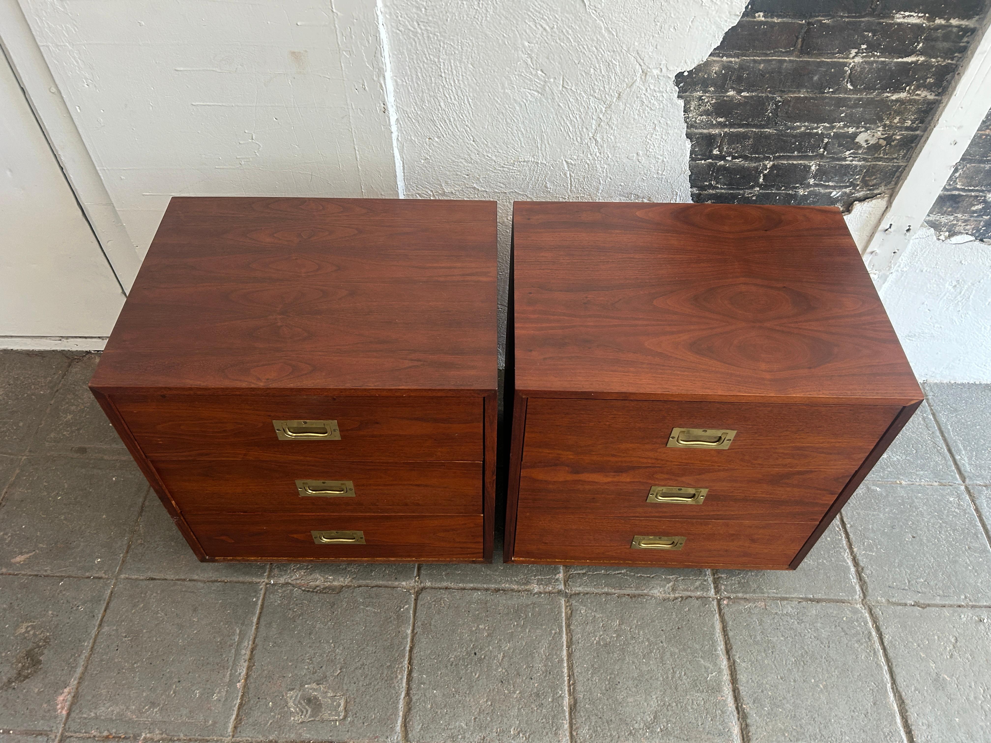 Mid century pair of 3 drawer walnut brass campaign style nightstands. Pair of small nightstands with 3 drawers campaign style with solid brass fold down handles. Each unit has a set of 4 small square legs. Good vintage condition made circa 1970 in