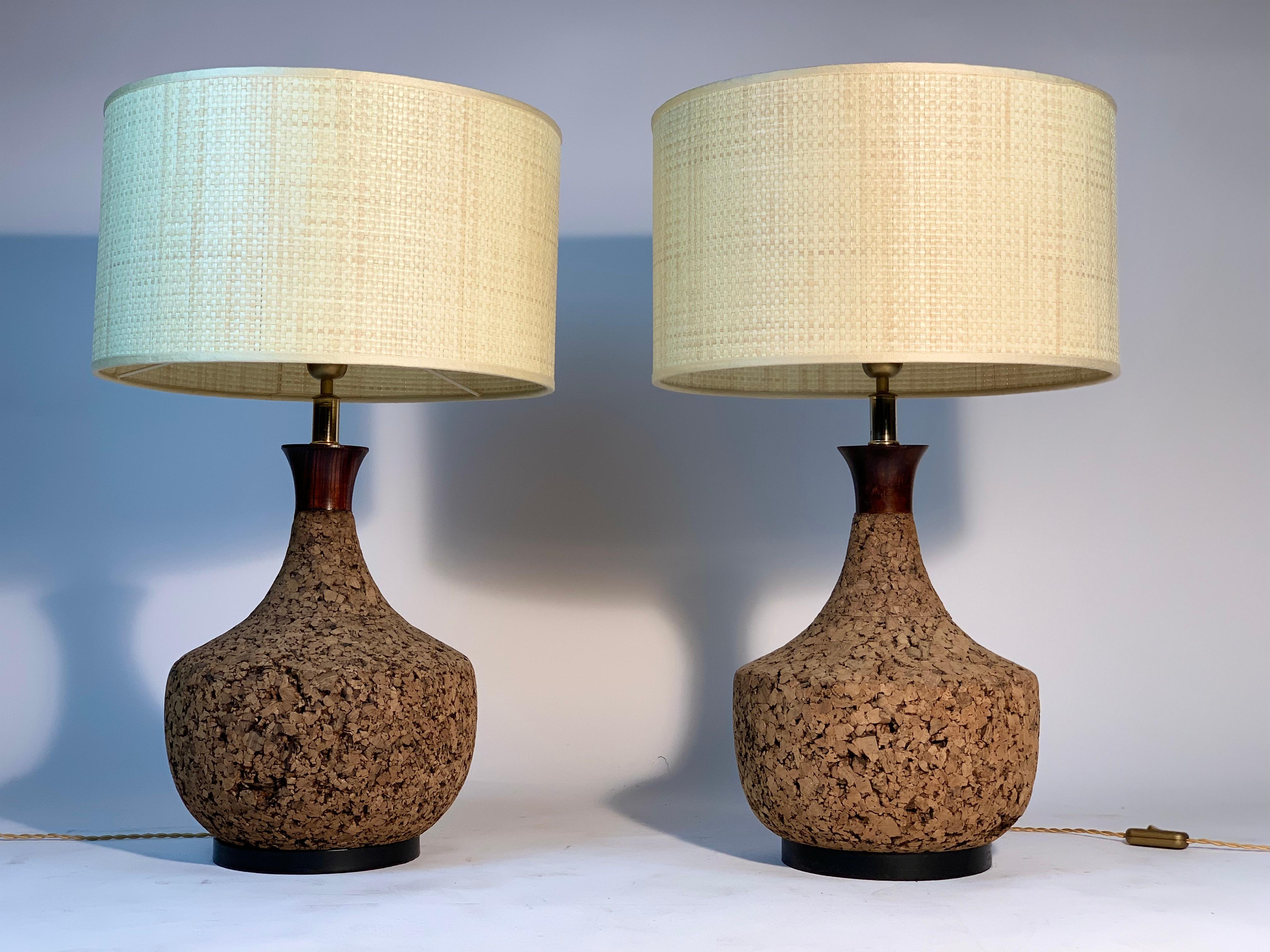 Pair of Italian table lamps in natural cork with dark lacquered metal base, in the shape of a pot-bellied bottle.
The collar at the top is made of wood. 
Italian production of the 1960s.
They feature a woven raffia lampshade.
The measurements in