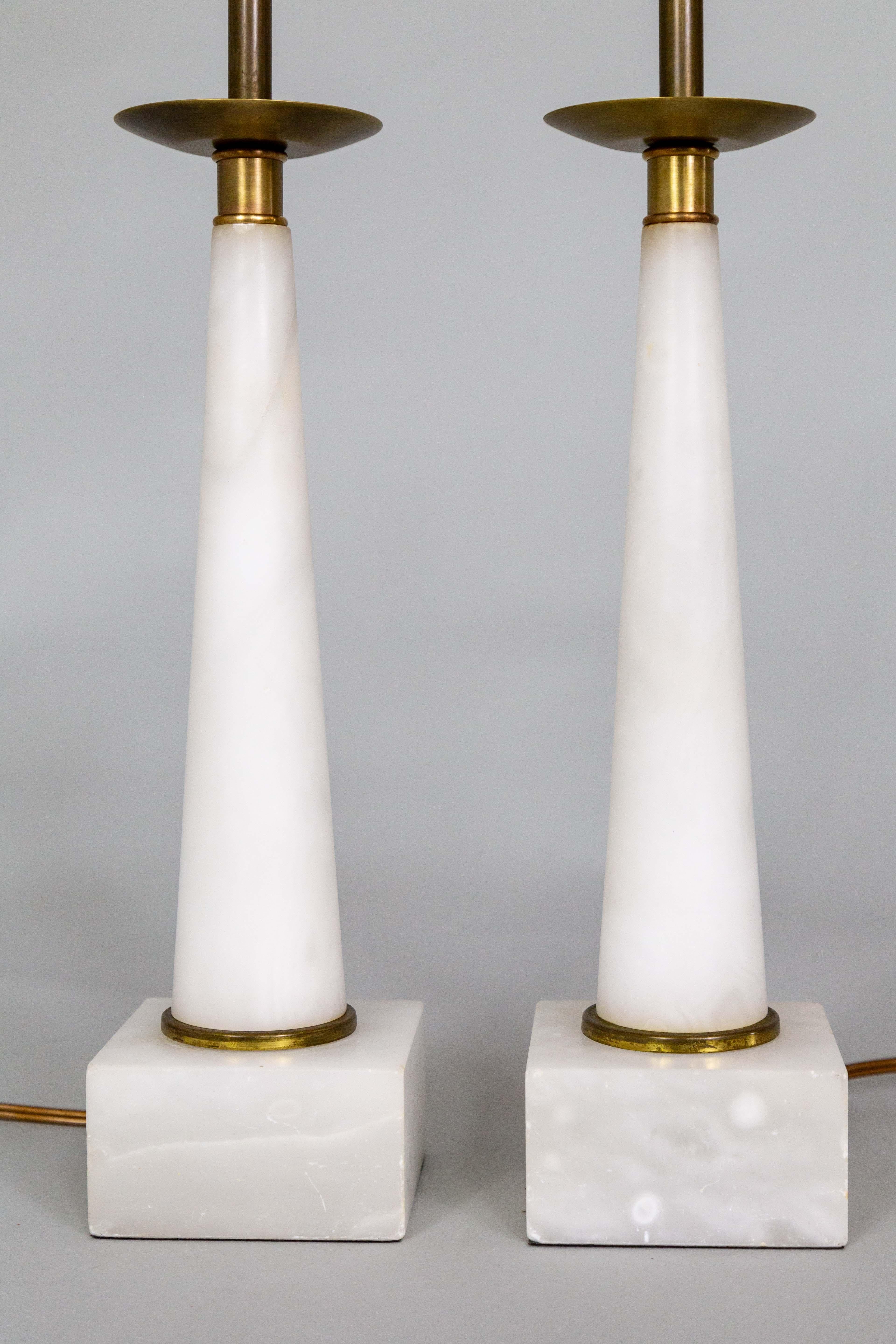 A pair of modern, slim candlestick lamps carved from alabaster as conical columns with square bases.  Made in the 1960s in a Tommi Parzinger style. Accented with dark, patinated brass necks and rims at the base. A versatile design that fits many
