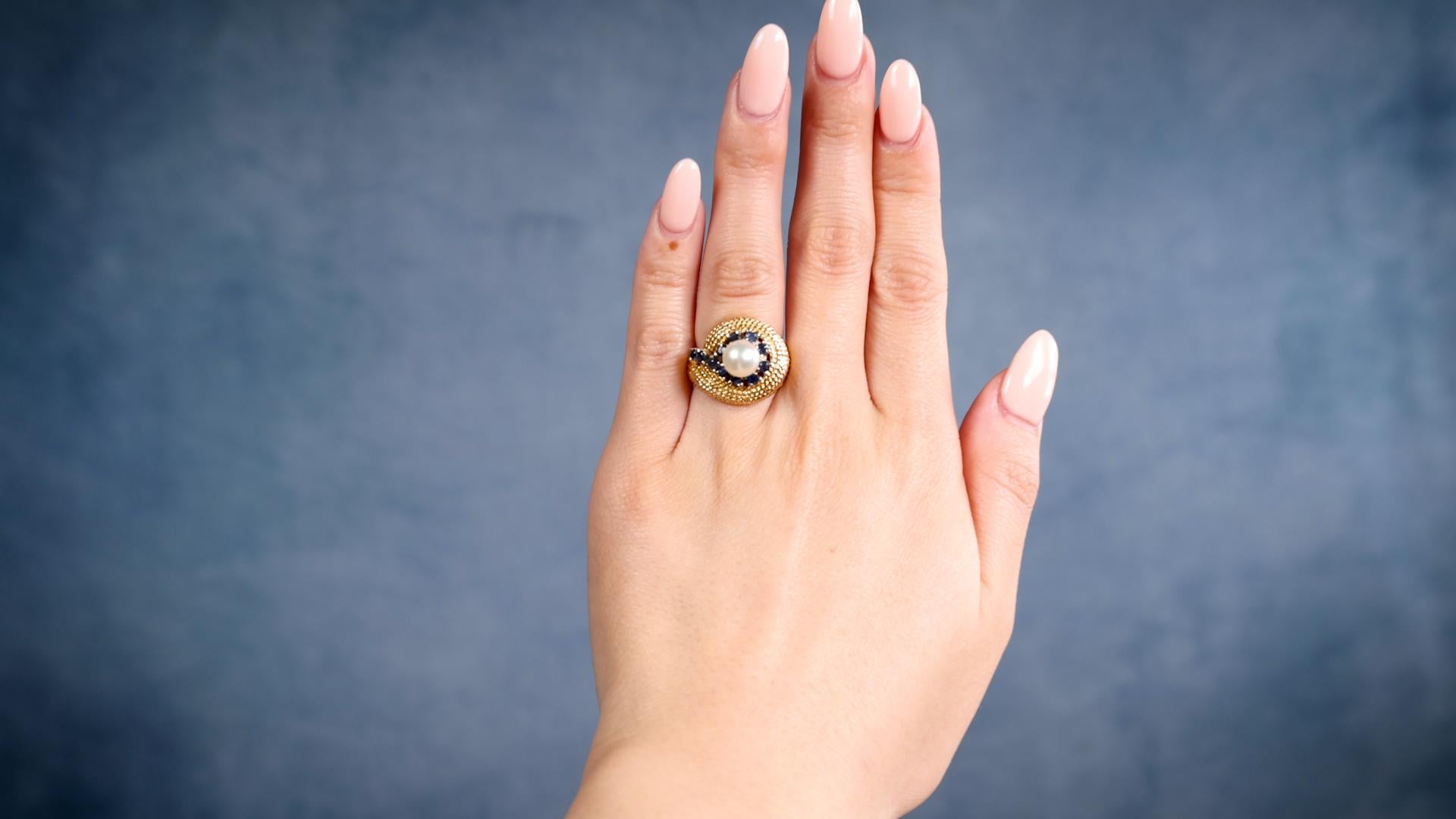 Ein Mid-Century Perle Saphir 14k Gelbgold Ring. Mit einer cremefarbenen Perle mit roséfarbenem Unterton, die etwa 6,90 Millimeter misst. Akzentuiert durch 11 runde Saphire mit einem Gesamtgewicht von etwa 0,25 Karat. Gefertigt aus 14 Karat Gelbgold