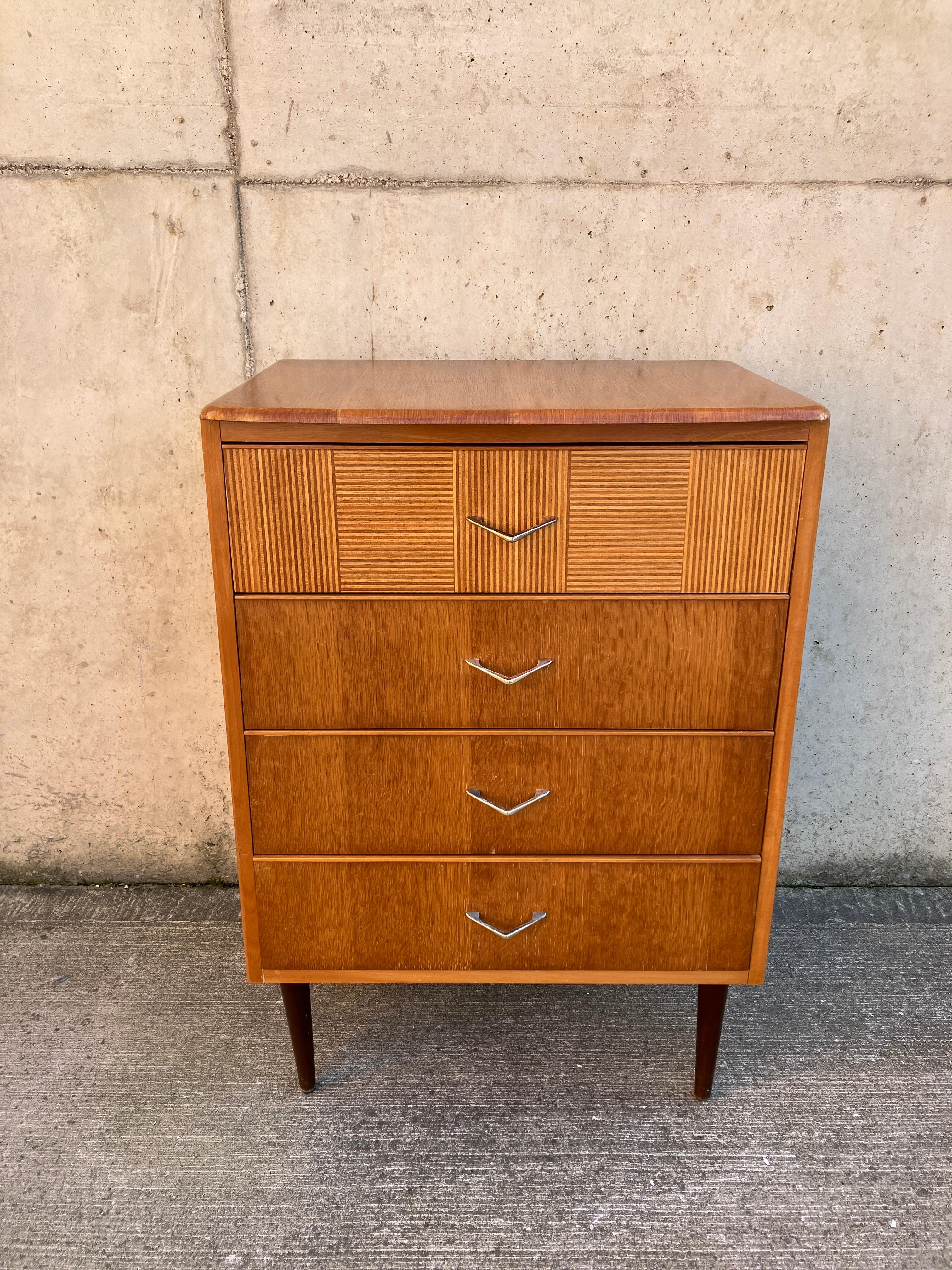 Eine kleine Kommode aus blondem Holz aus den 1950er Jahren. Es hat vier Schubladen mit goldenen Griffen und eine schön verzierte obere Schubladenfront. Es ist ein wahres Festival der Briten. In einer Schublade befindet sich ein roter Stempel/eine