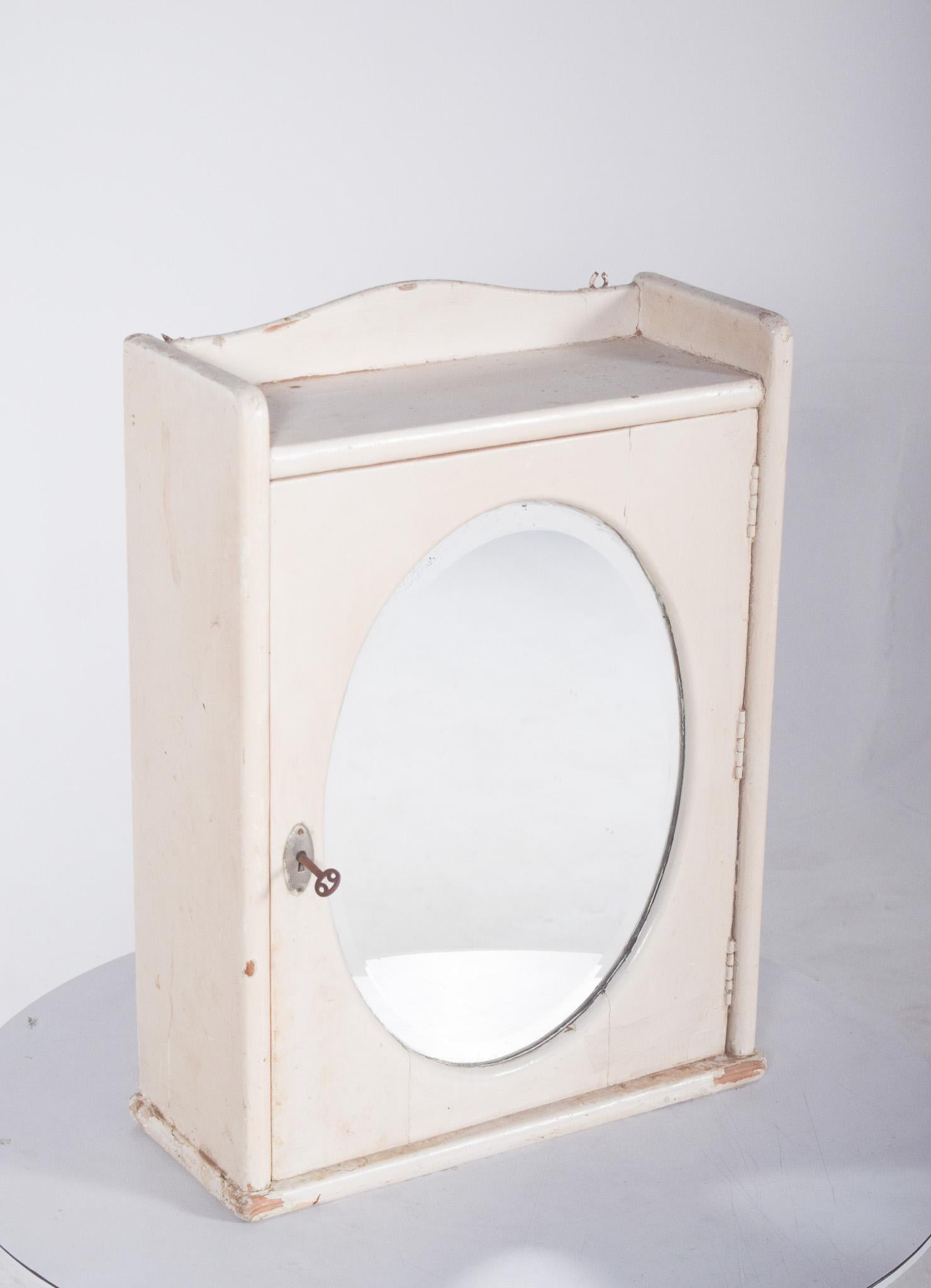 Pharmacy cabinet with oval mirror on the door. In white painted wood, it has a door with an oval mirror and a key, which suggests that it could be used for storing personal items or medicines. Inside, the cabinet has small shelves and vertical