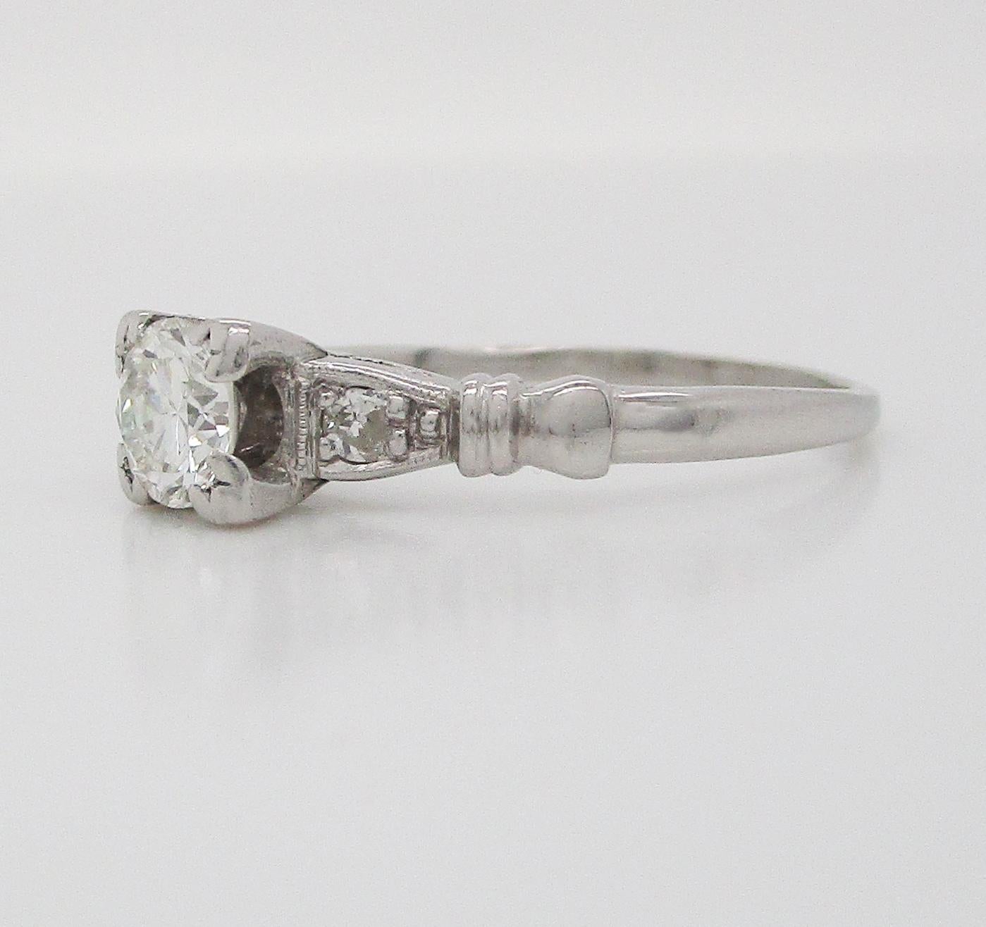 This is a stunning mid-century diamond engagement ring in platinum with lovely engraved shoulders. The ring features a diamond on each shoulder, framed by engraved details and accented by a fluted element on each shoulder. This ring is full of
