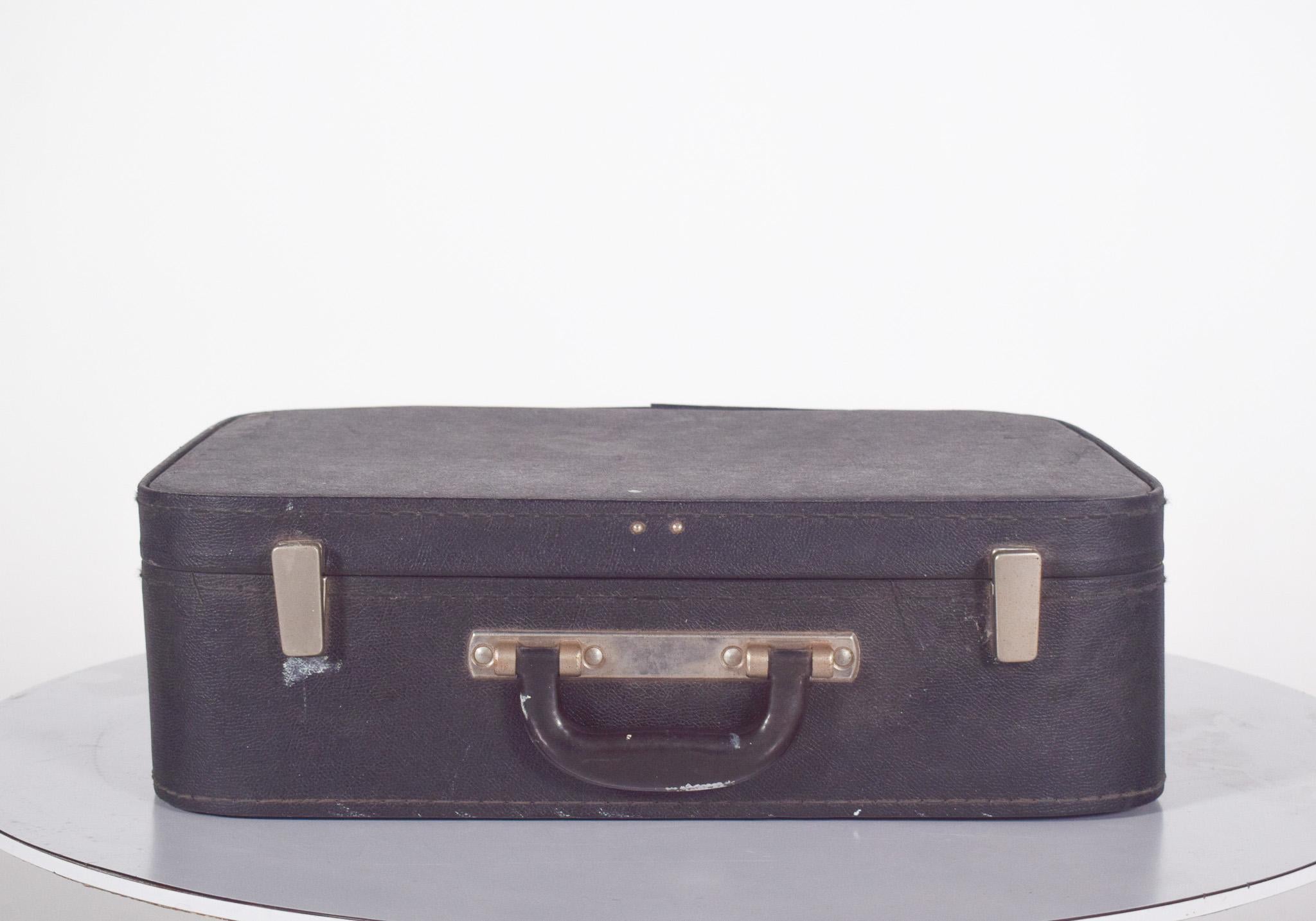Une valise ancienne. Recouvert à l'extérieur d'un mat�ériau synthétique noir texturé, avec des coins doucement arrondis. Il est doté de simples fermoirs métalliques sur les côtés et d'une poignée centrale renforcée en plastique noir avec des ferrures