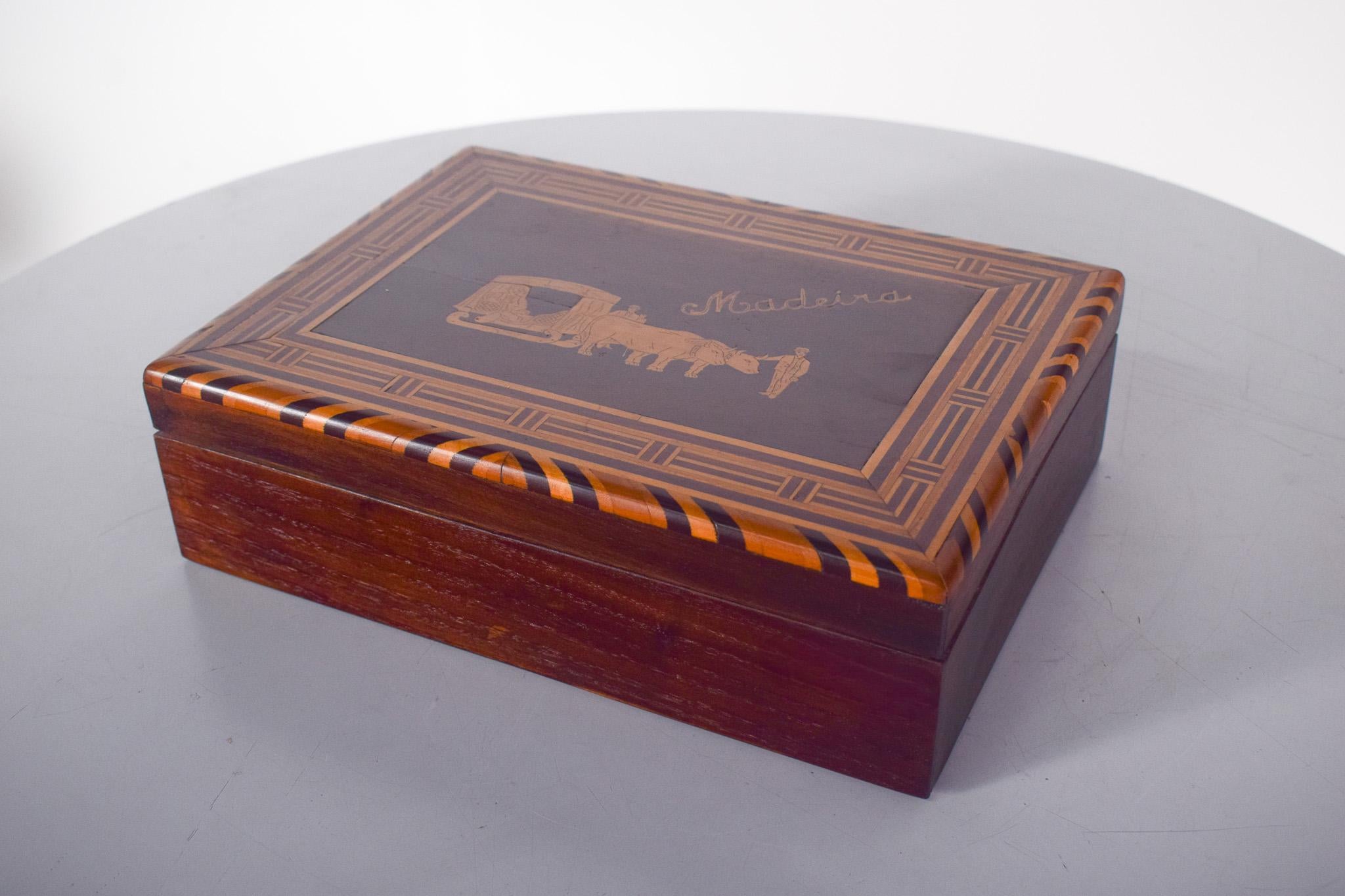 Scatola da cucito in legno. Il coperchio è incernierato, con intarsi e al centro l'immagine di un carro di buoi con conducente, tipico dell'isola di Madeira, decorato tutt'intorno con un motivo geometrico. Una volta aperto, rivela diversi piccoli