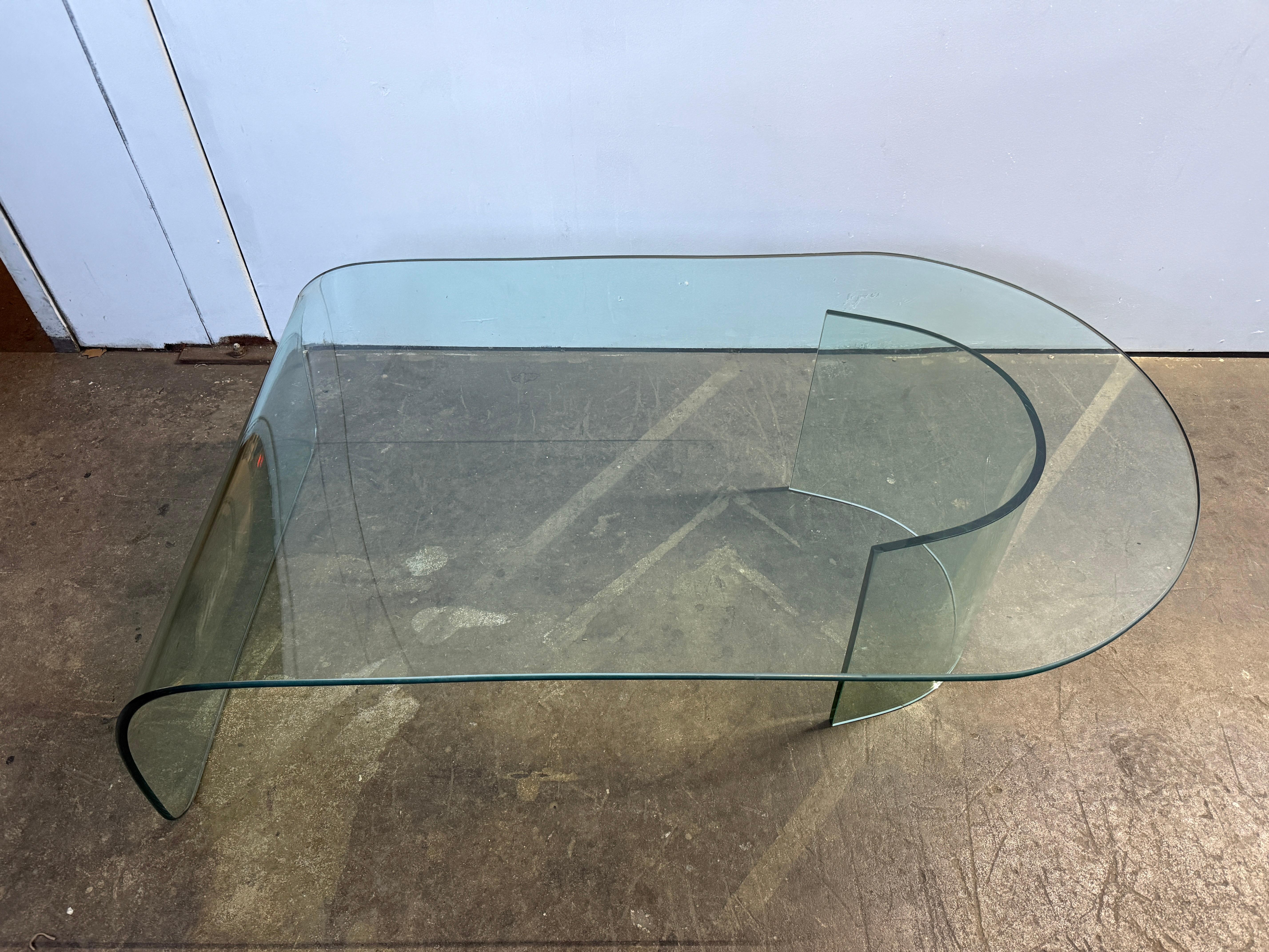 Mesa de centro posmoderna con tablero en cascada que descansa sobre un soporte curvado en forma de C. El cristal está muy limpio, sin astillas ni arañazos importantes. Puede tener un ligero desgaste de uso normal en la superficie. Bordes pulidos
