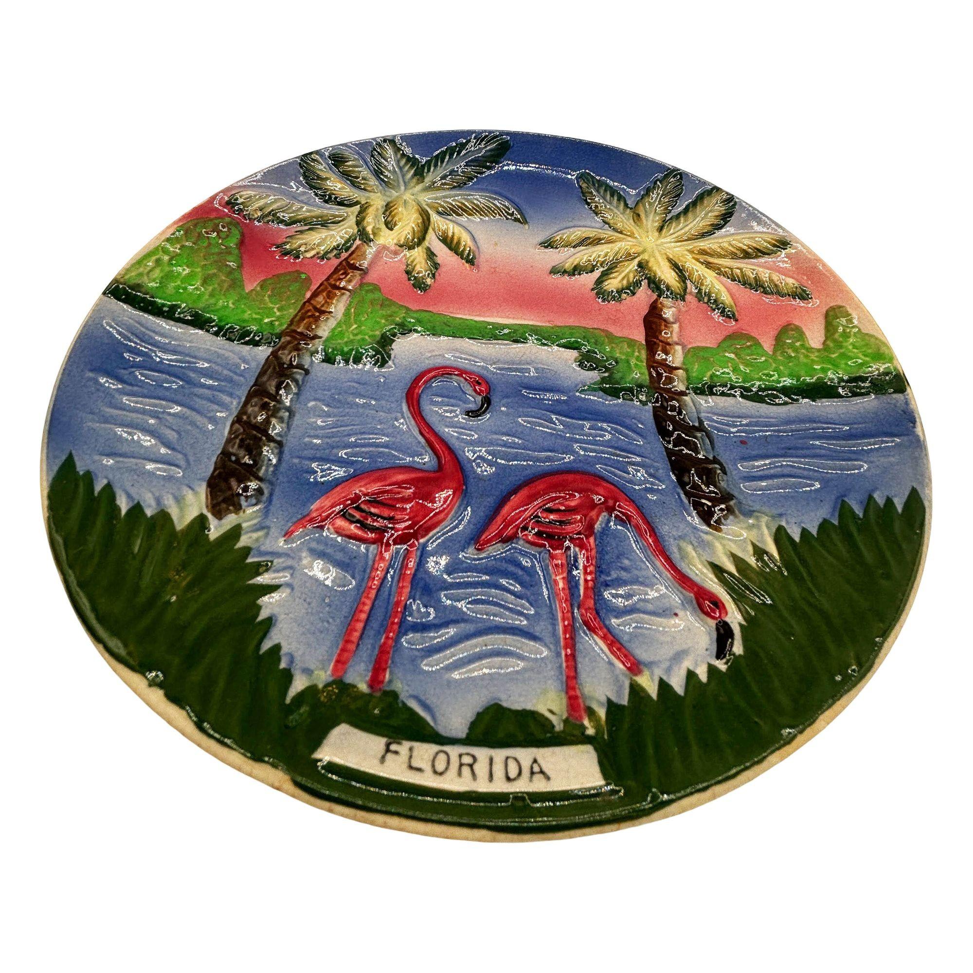 Plato decorativo de pared de cerámica de mediados de siglo con motivos de flamencos rosas y verdes. La placa de pared muestra una pareja de flamencos rosas bellamente detallados en una playa al atardecer, con el estado de Florida escrito en la parte