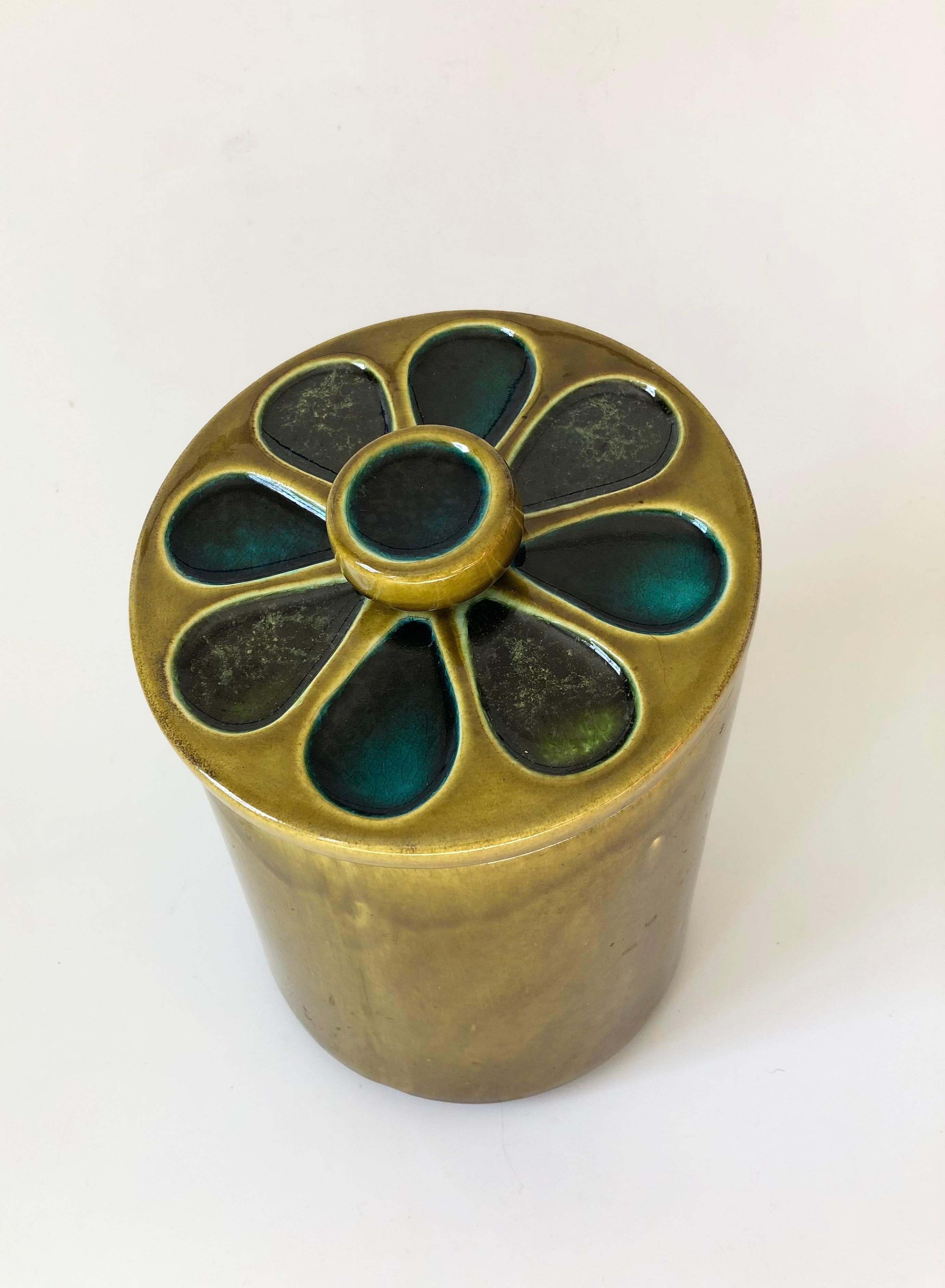 A mid century pottery kitchen canister. Flower design to the lid with a slightly angled top. Olive green glaze with blue petals to the flower on the lid. Made in California.


