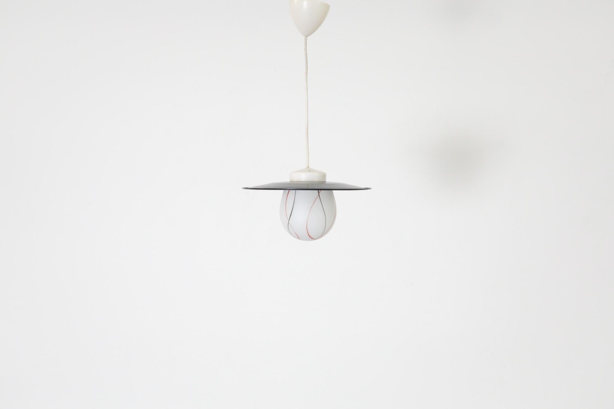 Pendente da soffitto Philips di metà secolo con paralume rotondo smaltato nero e diffusore grafico in vetro lattimo stampato rosso e nero. In condizioni originali, con un'usura coerente con l'età e l'uso. L'altezza complessiva, compreso il Corde, è
