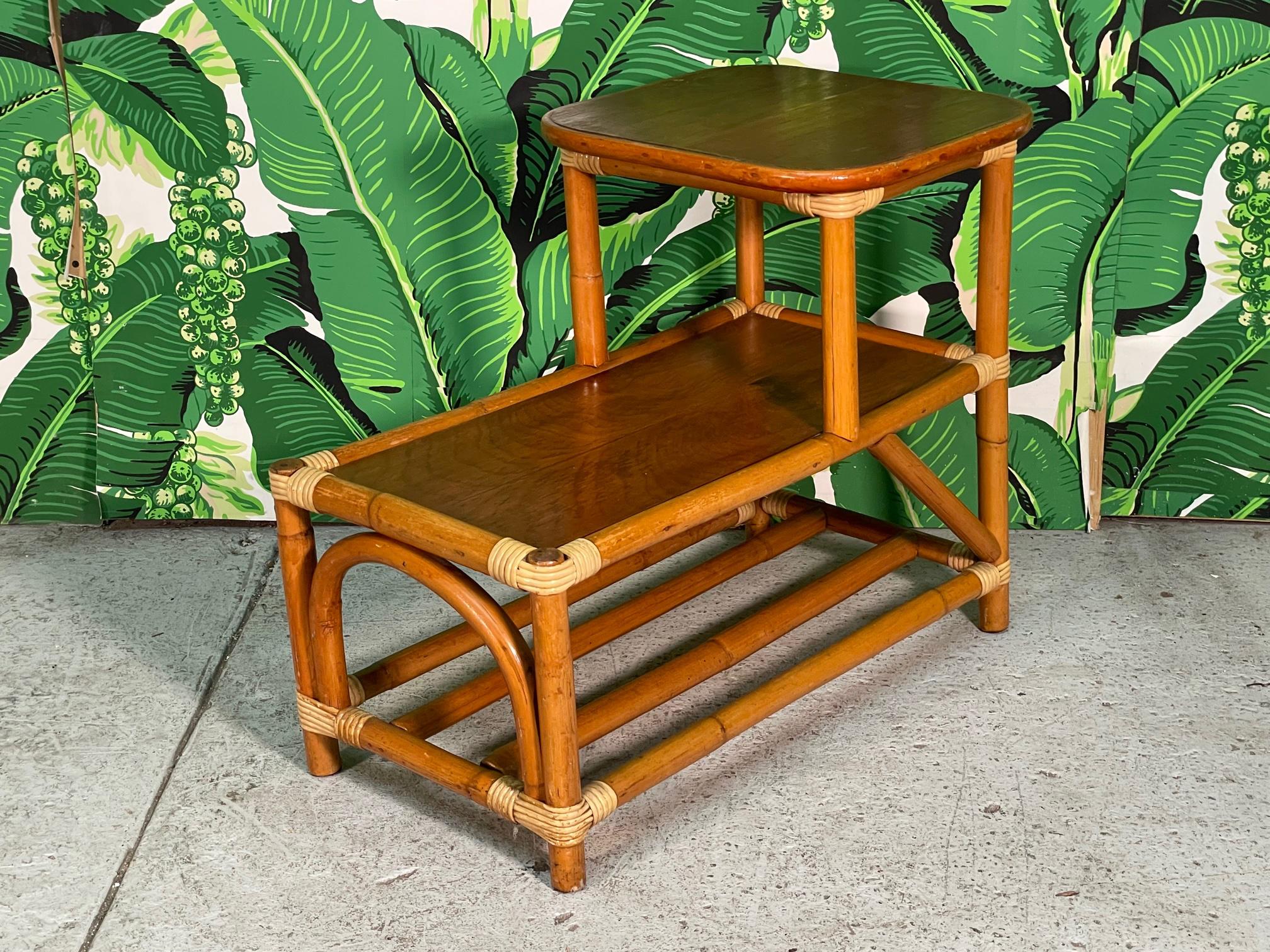 Midcentury Rattan two-tiered side table features mohagany tops and contrasting strapping. Horizontal poles between stretchers form lower shelf. Good condition with minor imperfections consistent with age. May exhibit scuffs, marks, or wear, see