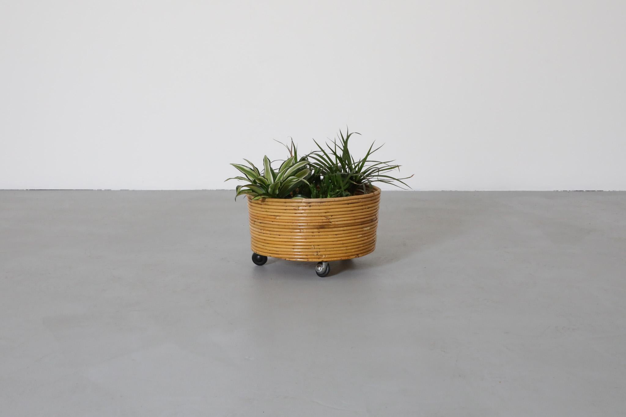Mid-Century round bamboo wrapped plant stand with round chrome and black wheels. A light weight indoor planter ideal for adding a stylish and organic touch to a space. In original condition with visible wear consistent with its age and use. Plants