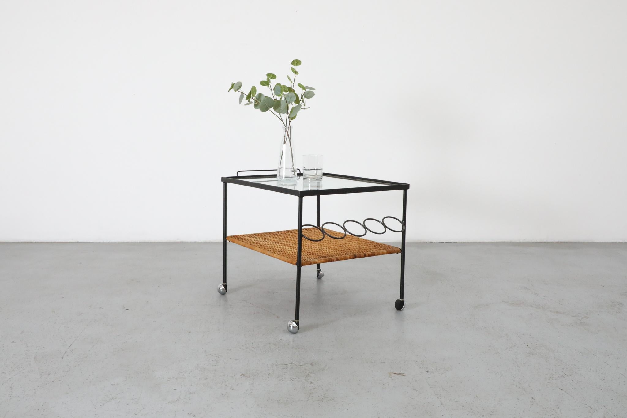 Rare Mid-Century rolling bar cart in the style of acclaimed French designer Mathieu Matégot. Designed in the 1950s, this elegant and functional piece features a clear inset glass top, a woven rattan lower shelf, and a dedicated bottle storage