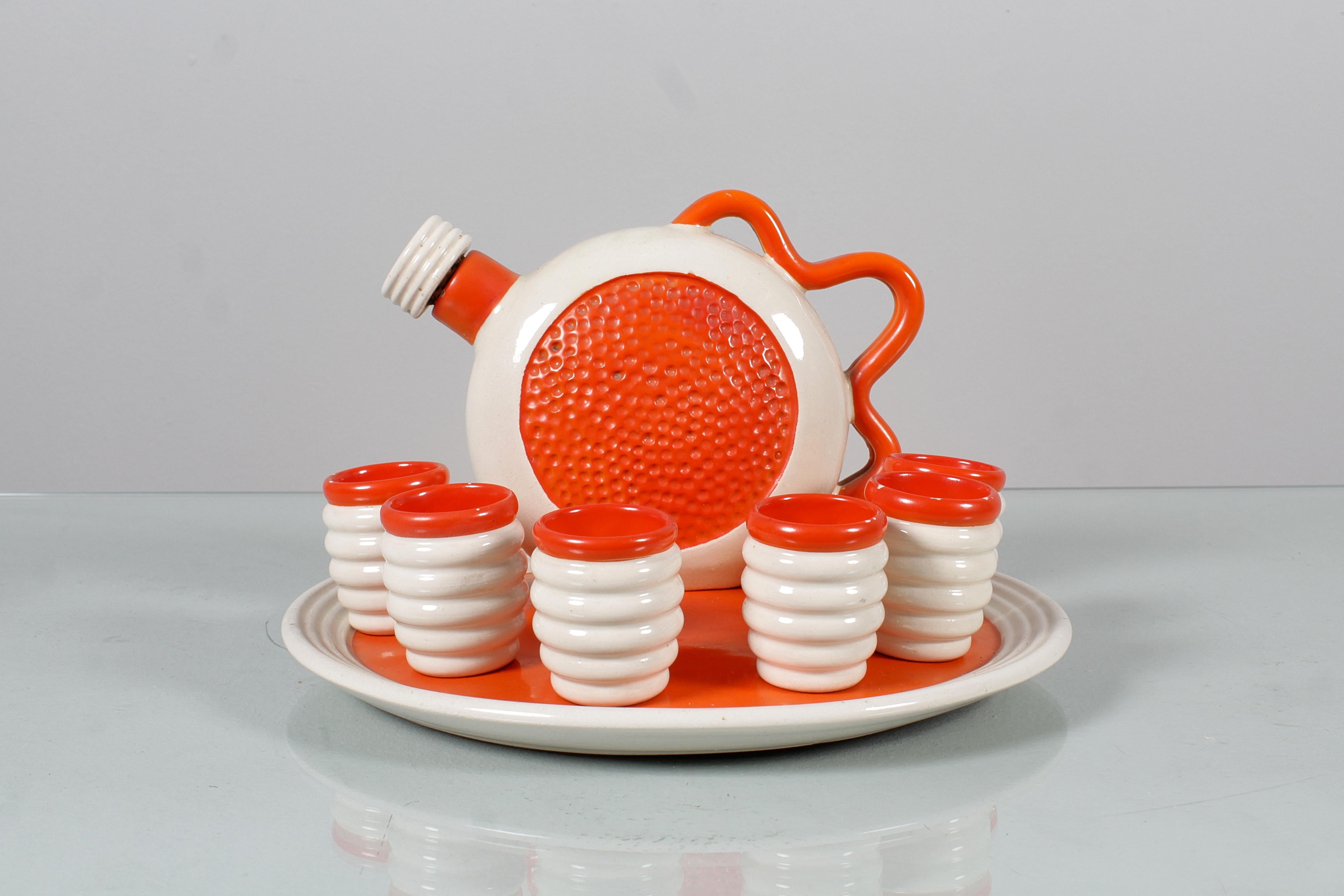 Sehr originelles Likörset aus gearbeiteter und glasierter Keramik in Weiß- und Orangetönen, bestehend aus einer Flasche mit Henkel, sechs Gläsern und einem runden Tablett. Das Set ist auf dem Boden des Tabletts und der Flasche signiert: 