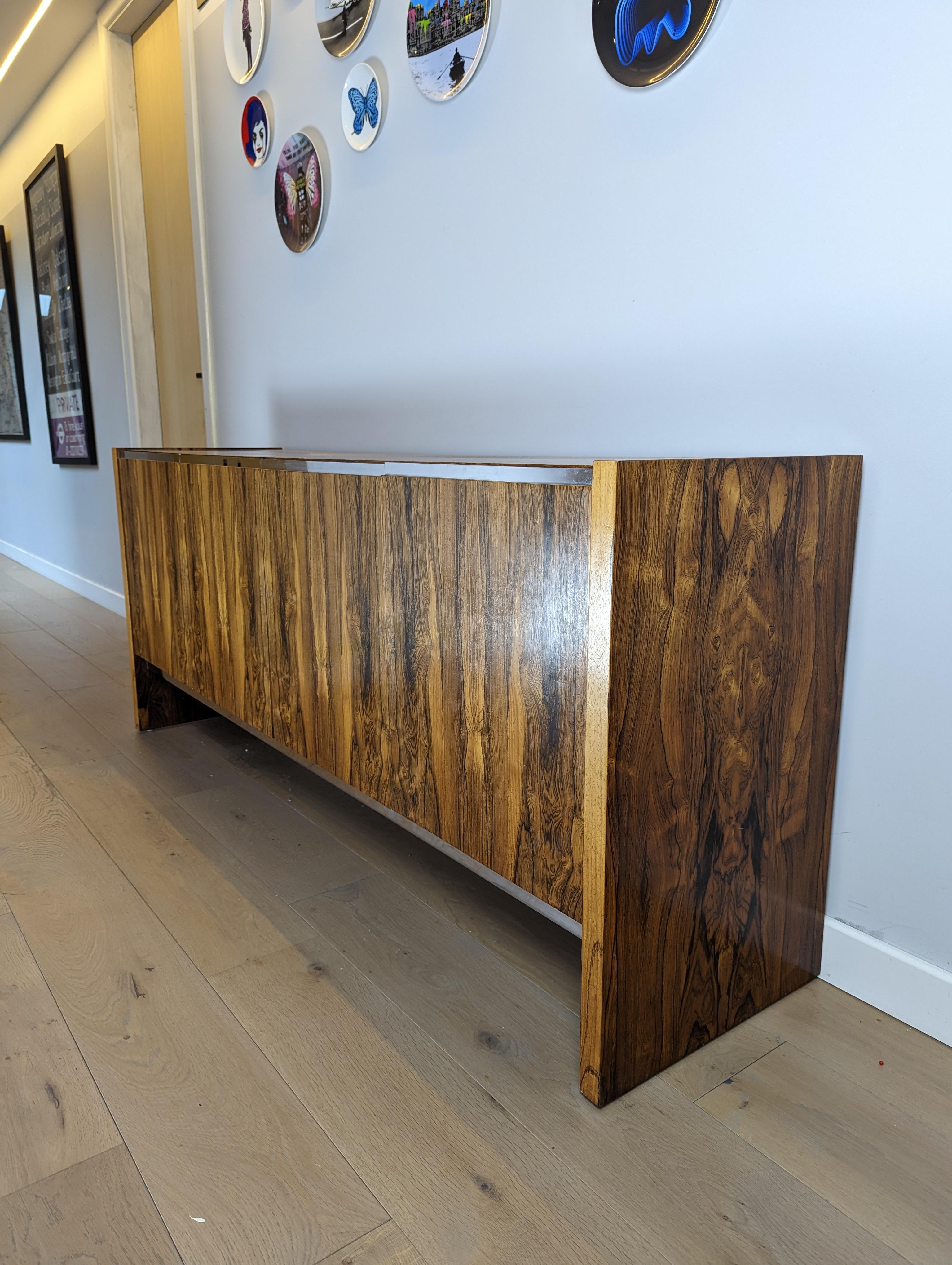An absolutely stunning vintage sideboard in rosewood with chrome accents, made by Merrow Associates. This was designed by Richard Young; it dates from the 1960-70’s.

The quality is outstanding, this has gorgeous rosewood grain patterns throughout
