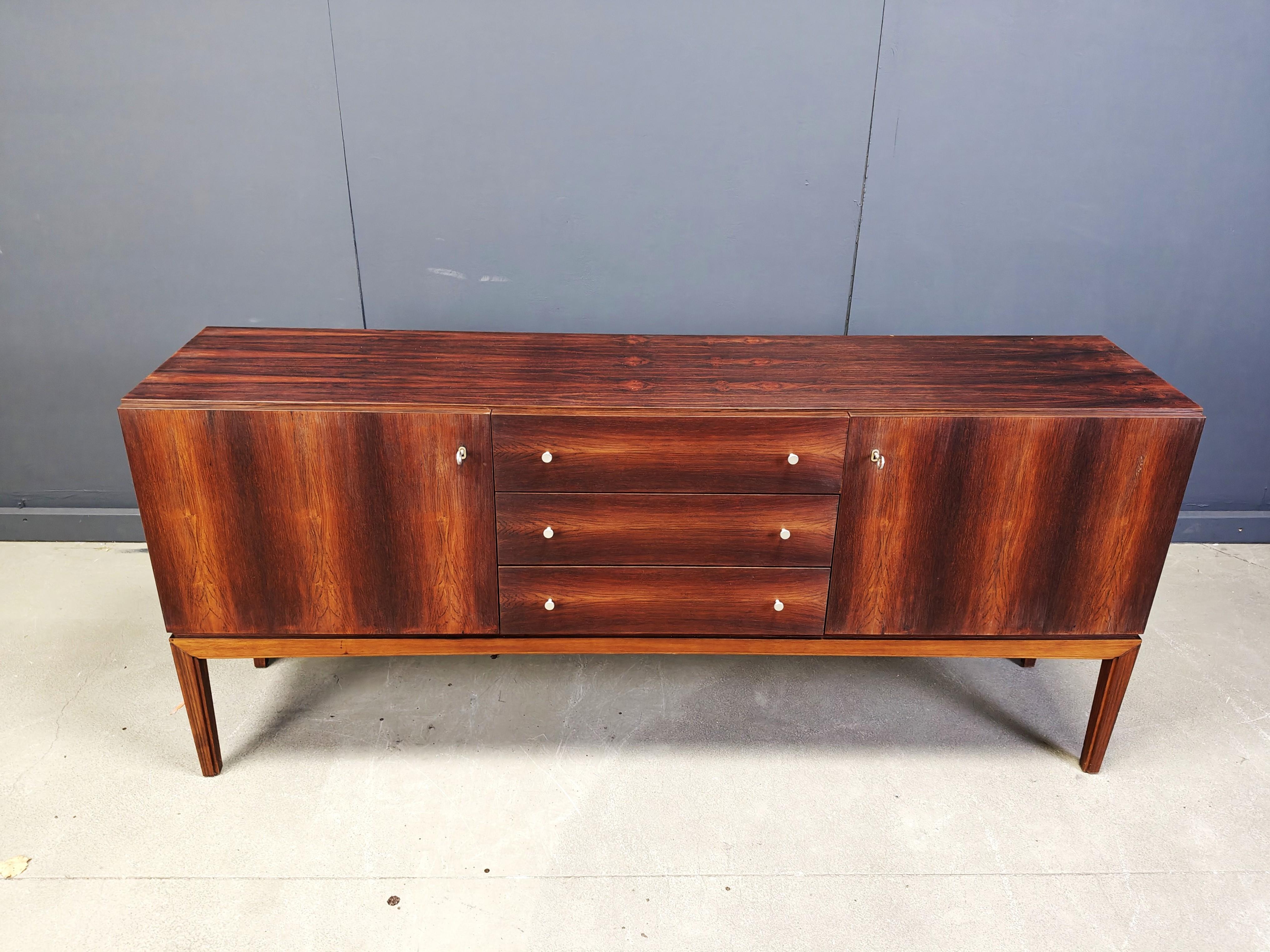 Credenza di metà secolo impiallacciata in palissandro con tre cassetti centrali e due ante.

Bellissimi pannelli impiallacciati in palissandro che mostrano una splendida venatura naturale.

La credenza, piccola ma funzionale, si integra facilmente