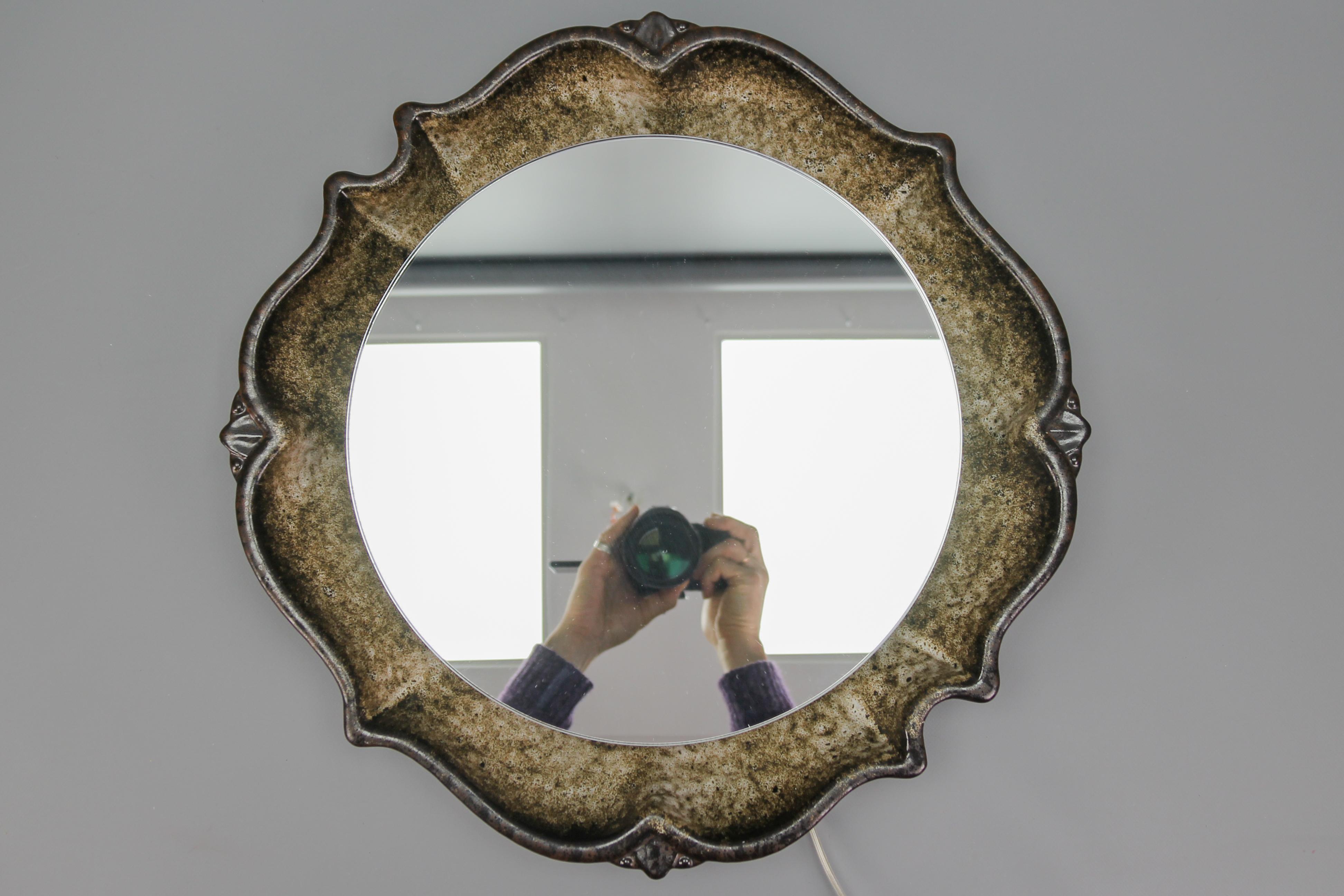 The high-quality glazed ceramic wall mirror was manufactured in Germany in the 1970s, in the Fat Lava Art Pottery period. The gracefully shaped ceramic frame shows beautiful warm brown tones. Centered with a round mirror with a backlight that