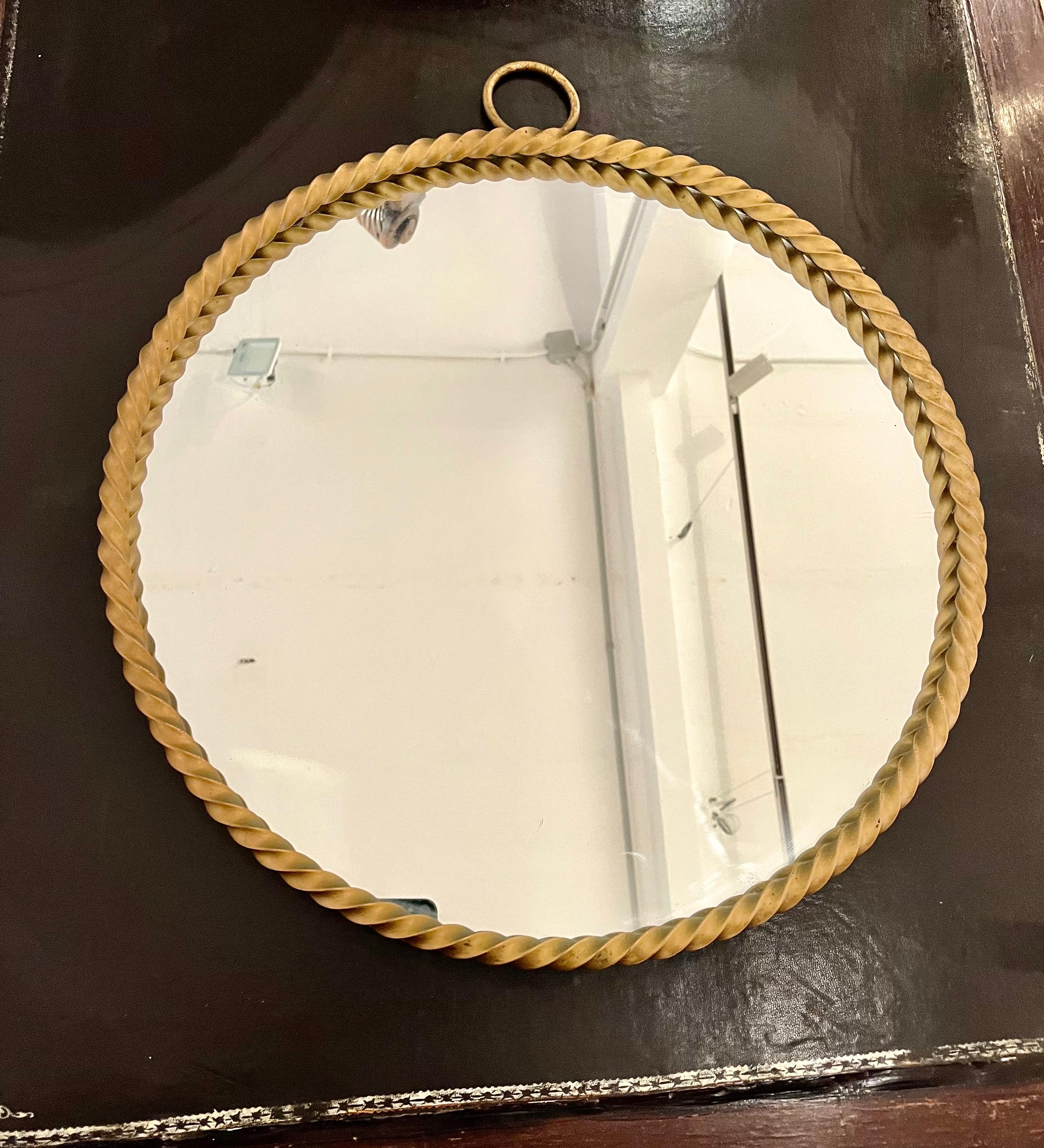 Uno specchio circolare circondato da una cornice in metallo dorato. Il metallo è stato attorcigliato per creare l'aspetto di una corda. In cima c'è un anello. La firma del designer è visibile sul retro, che porta la firma della stilista, Chaty
