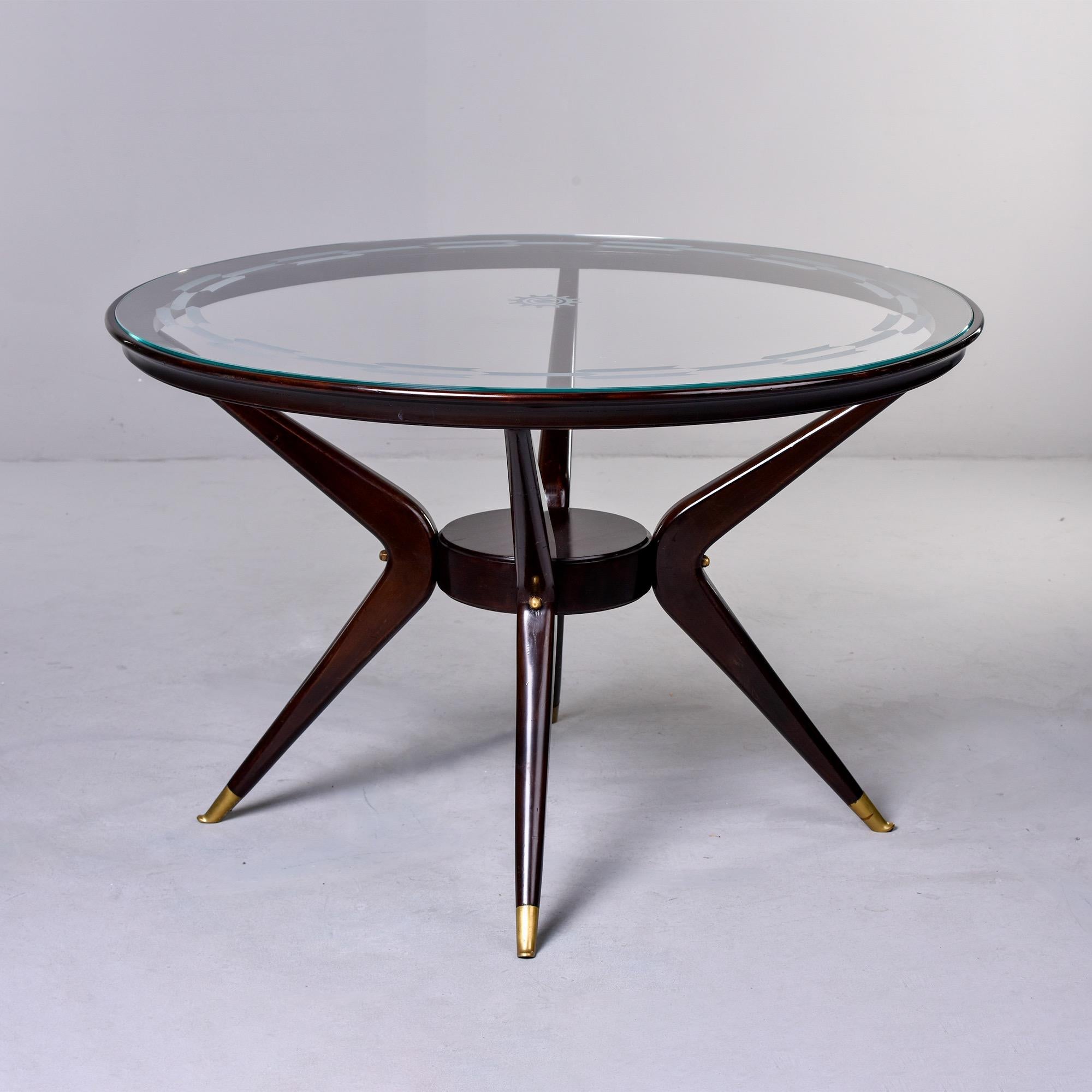 Circa 1970s Italian round table is just under 48” diameter and has a dark stained four leg wood frame with splayed and tapered brass capped legs. Inset glass top has a decorative etched border and small sunburst or gear wheel in center. Versatile