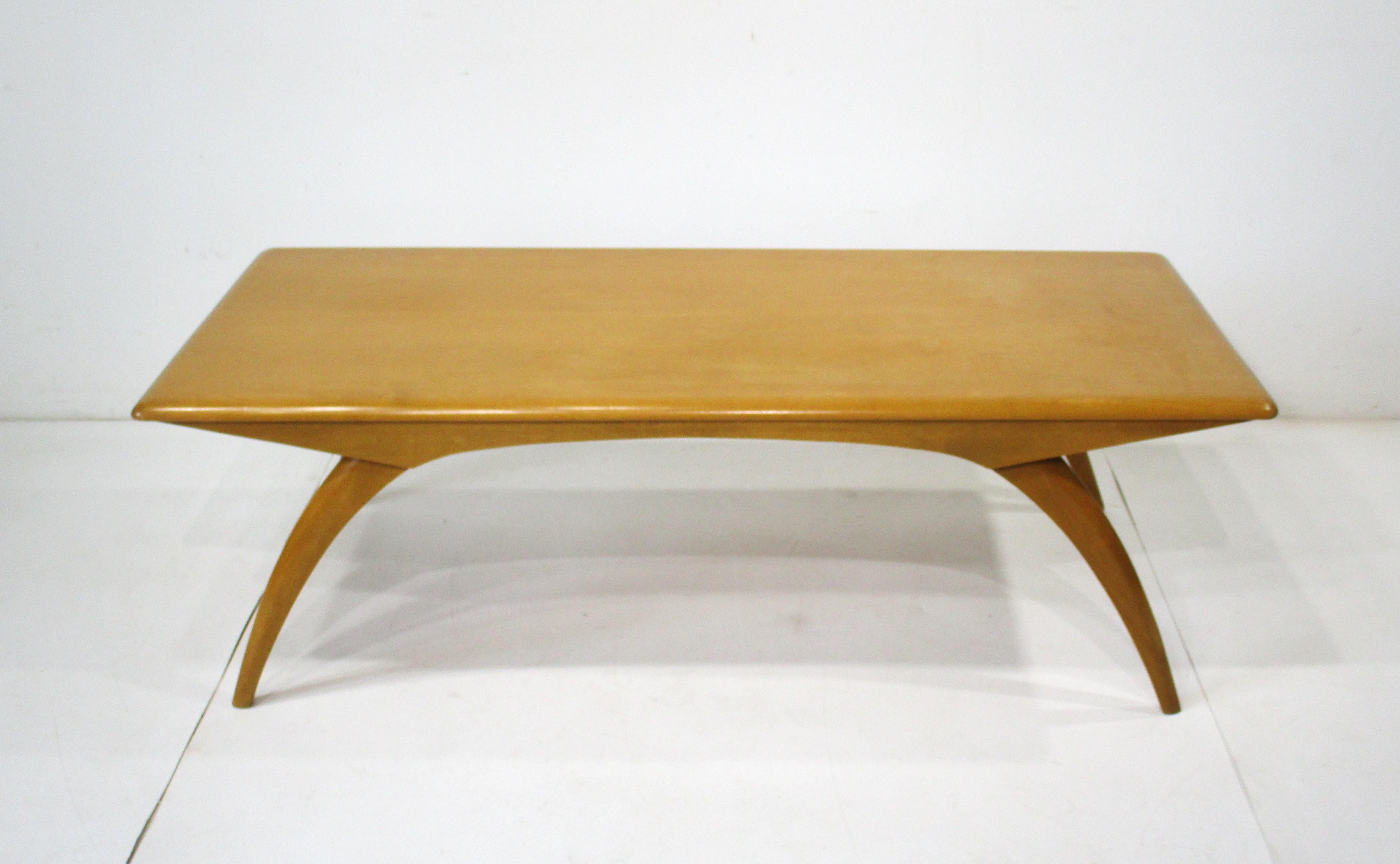 A Mid Century saber leg large cocktail table in solid maple wood with a wheat toned finish . Designed by Leo Jiranek and Ernest Herrmann in the 1950's this very well crafted piece is manufactured by the Heywood Wakefield furniture company model # M