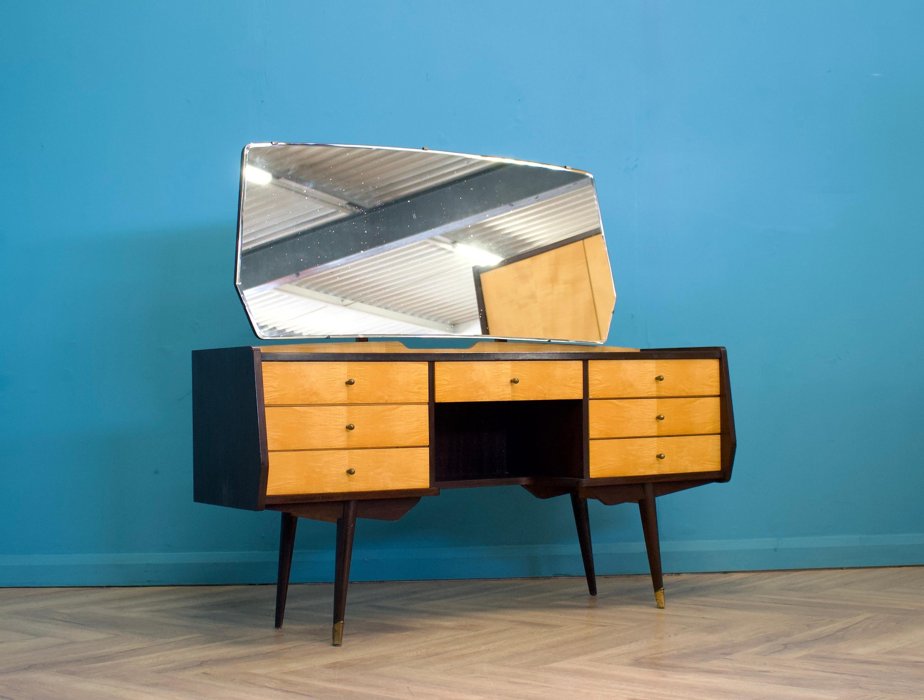 A mid-century birch and walnut dressing table from Times Furnishings, in the Italian style, circa 1960s.


It features a high gloss finish on the doors and splayed legs finished with brass details.


The dressing table includes seven drawers and a