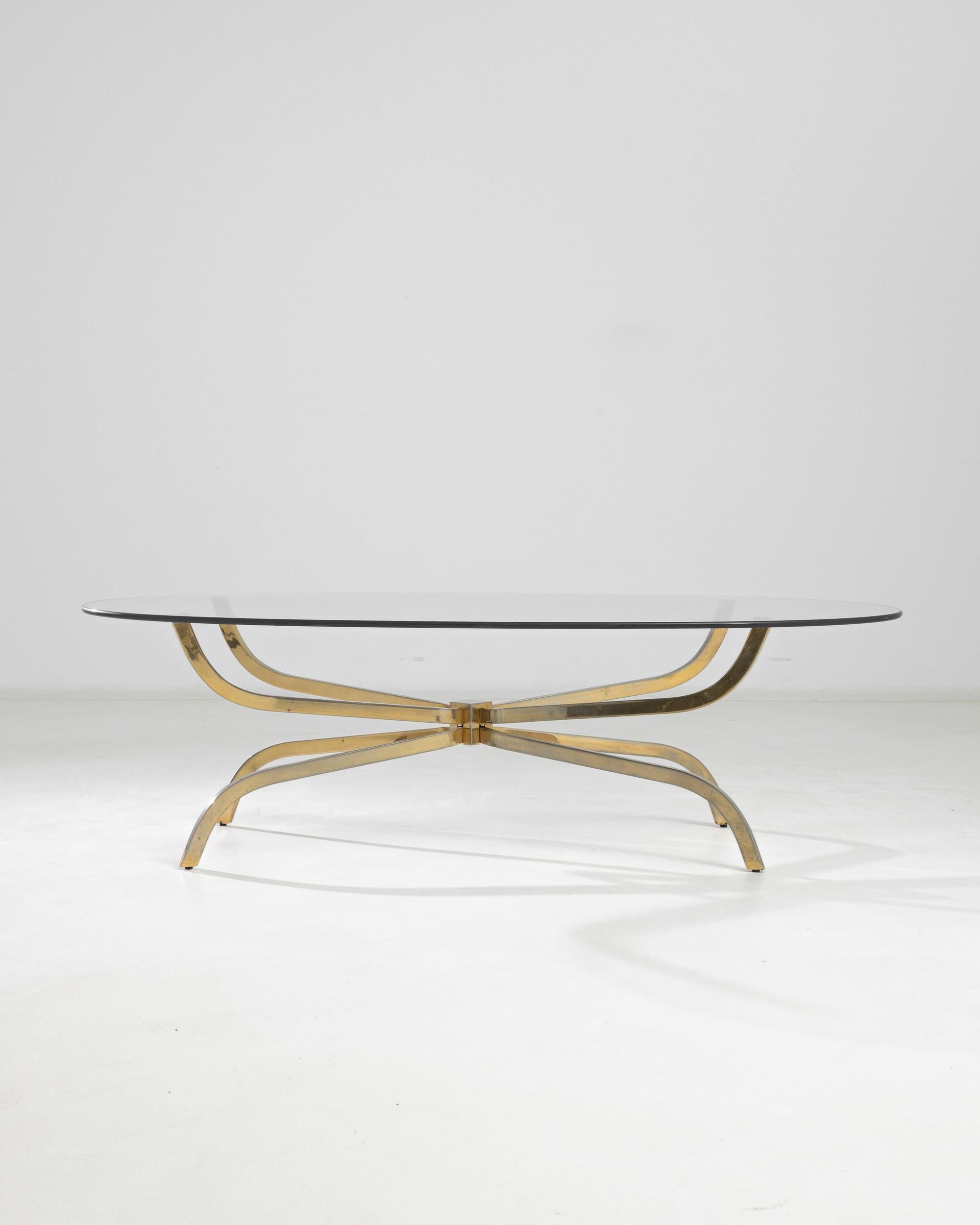 A 20th century brass coffee table produced in Scandinavia. An oval glass top rests on four bass feet, joined in the center like a minimalist floral bouquet. Sturdy yet delicate, the golden brass structure exhibits a light patina and the geometric