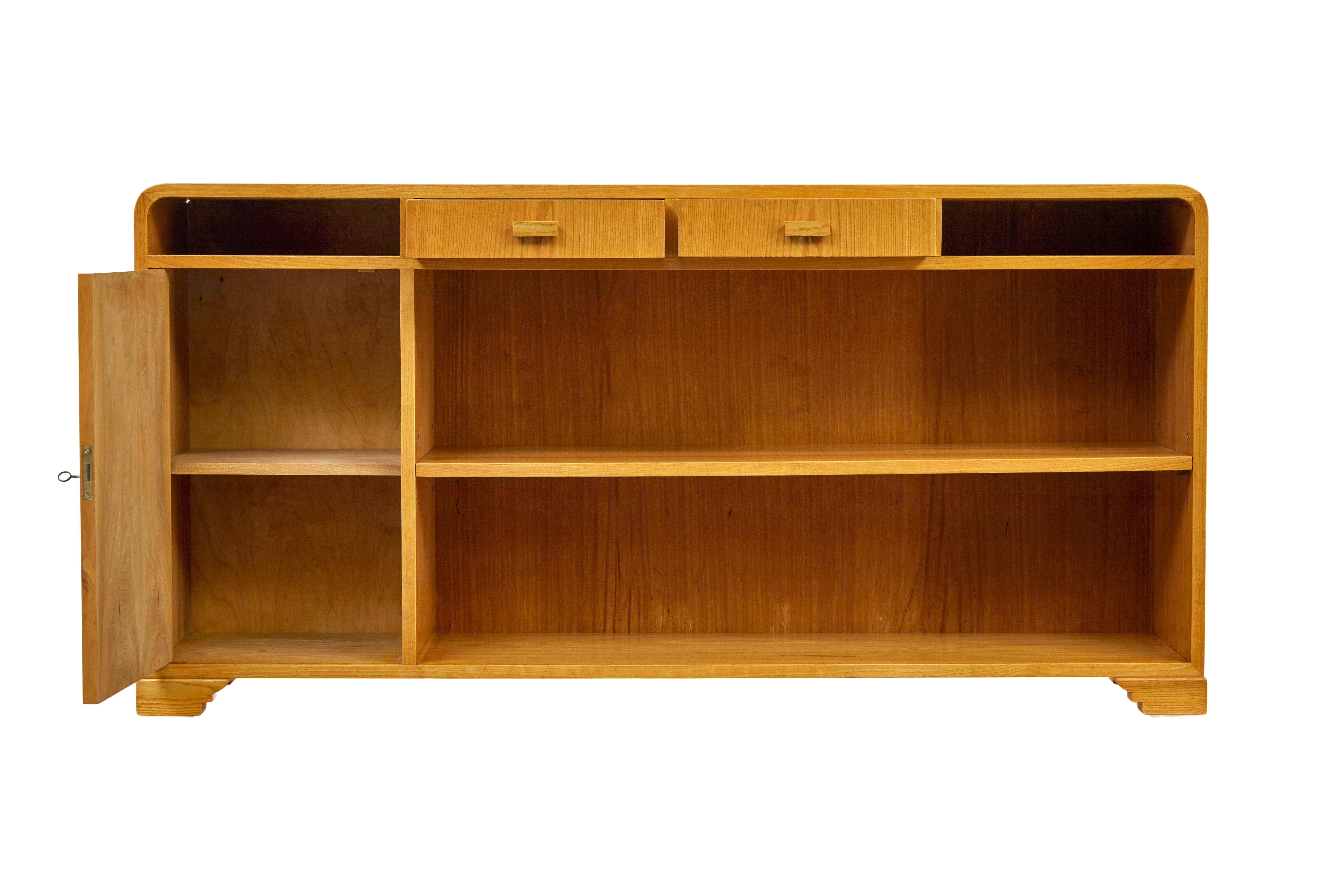 Mid century Scandinavian elm low open bookcase sideboard circa 1950.

Elegant piece of Swedish design with it's flowing lines and art deco inspiration.

Rich golden elm colour, featuring shaped corners.  2 central drawers below flanked either side