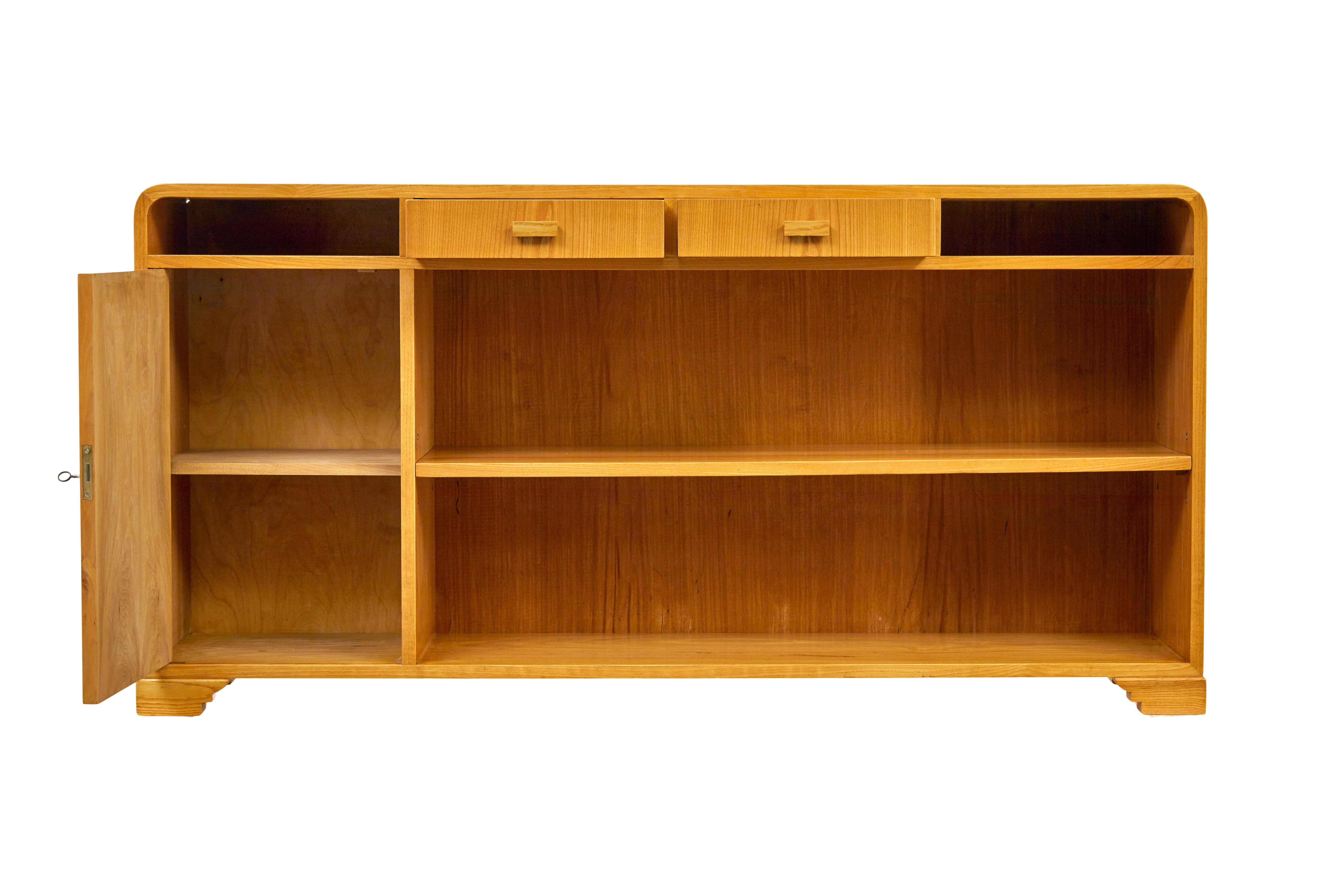 Mid century Scandinavian elm low open bookcase sideboard circa 1950.

Elegant piece of Swedish design with it's flowing lines and art deco inspiration.

Rich golden elm colour, featuring shaped corners.  2 central drawers below flanked either side