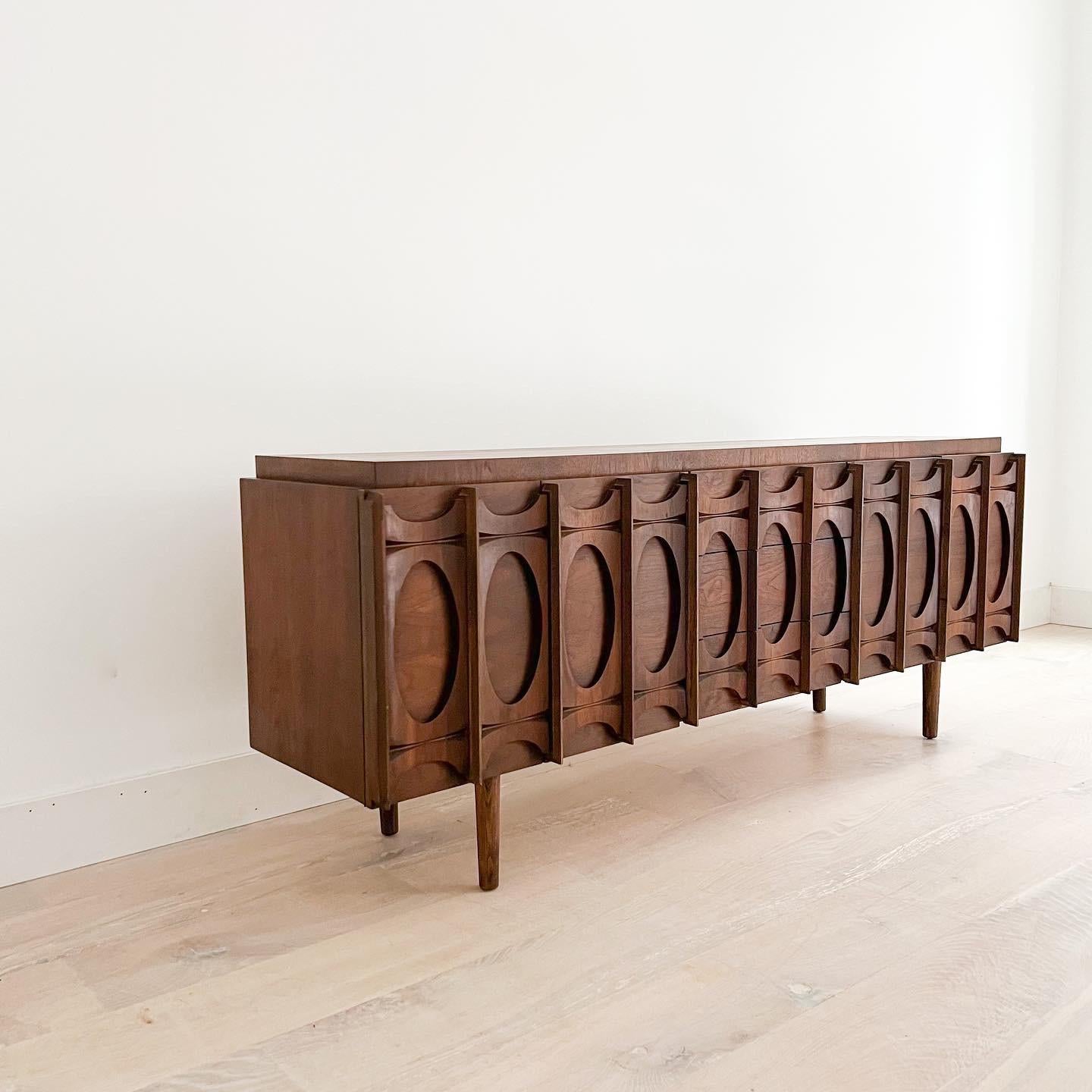 Mid century modern sculpted front walnut credenza on tapered legs by Tobago (made in Canada). The top has been sanded and restored. Some scuffing/scratching/small areas of veneer repair from age appropriate wear. Three middle drawers in the center
