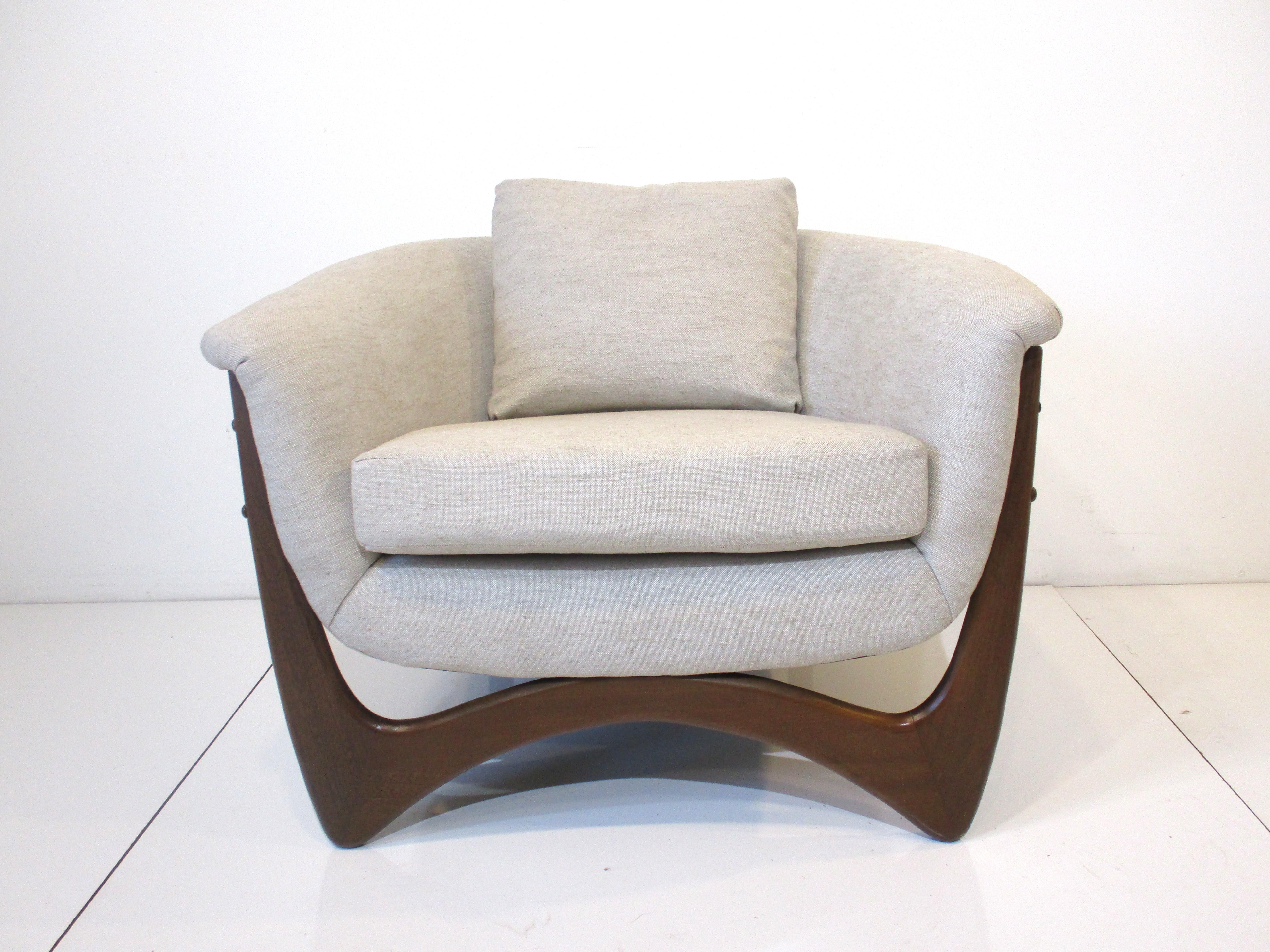A fully restored rounded backed lounge chair upholstered in a tightly woven contract fabric in a blend of sand, beige and cream. Designed for comfort with slightly tilted back and arms with loose back and bottom cushions. Having dark sculptural wood