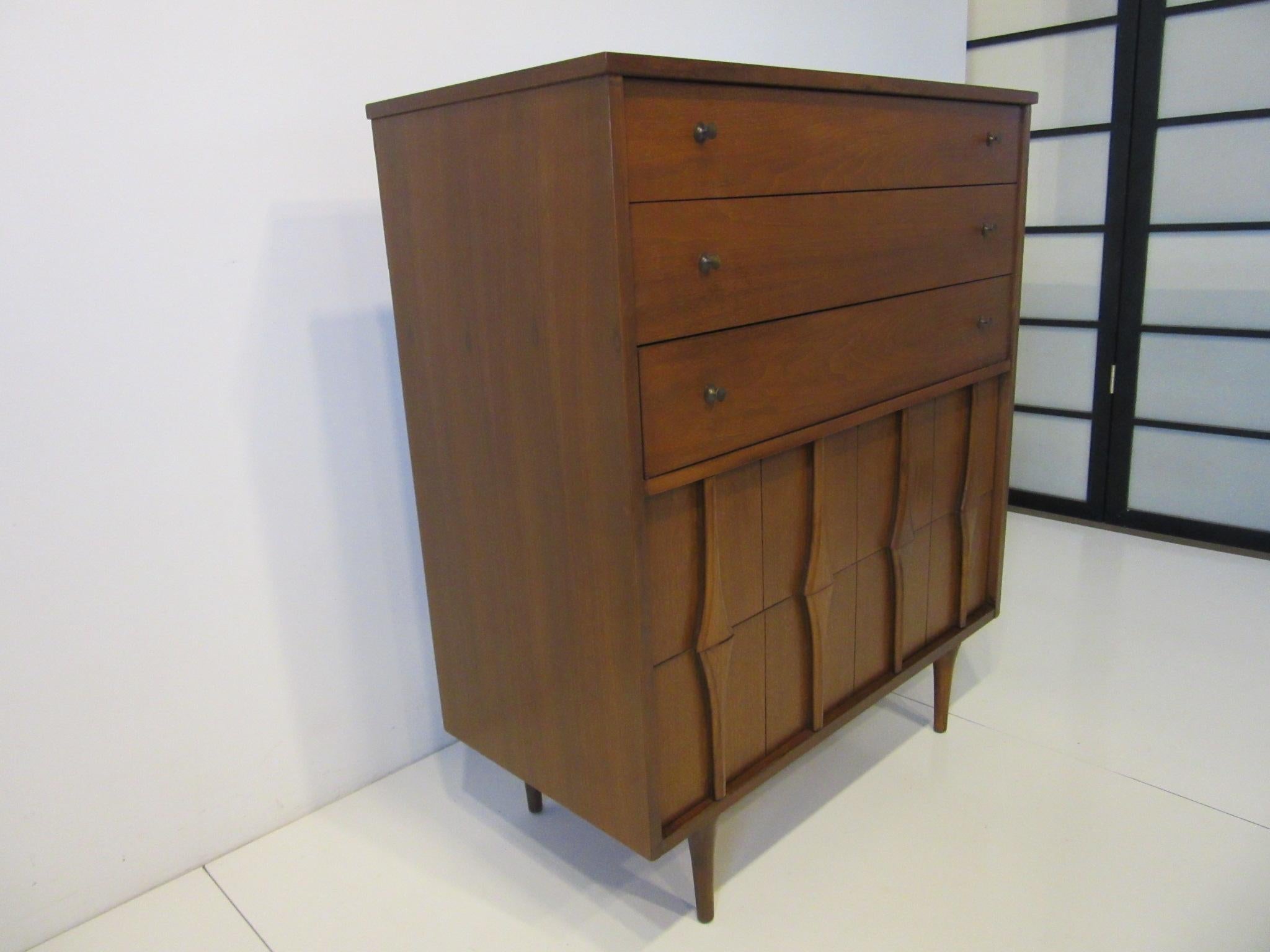 A tall walnut mid century dresser chest with three slim upper drawers having bronze toned pulls and two larger lower drawers with sculptural design fronts. Well made sitting on conical legs and with the walnut drawer details this piece will make a