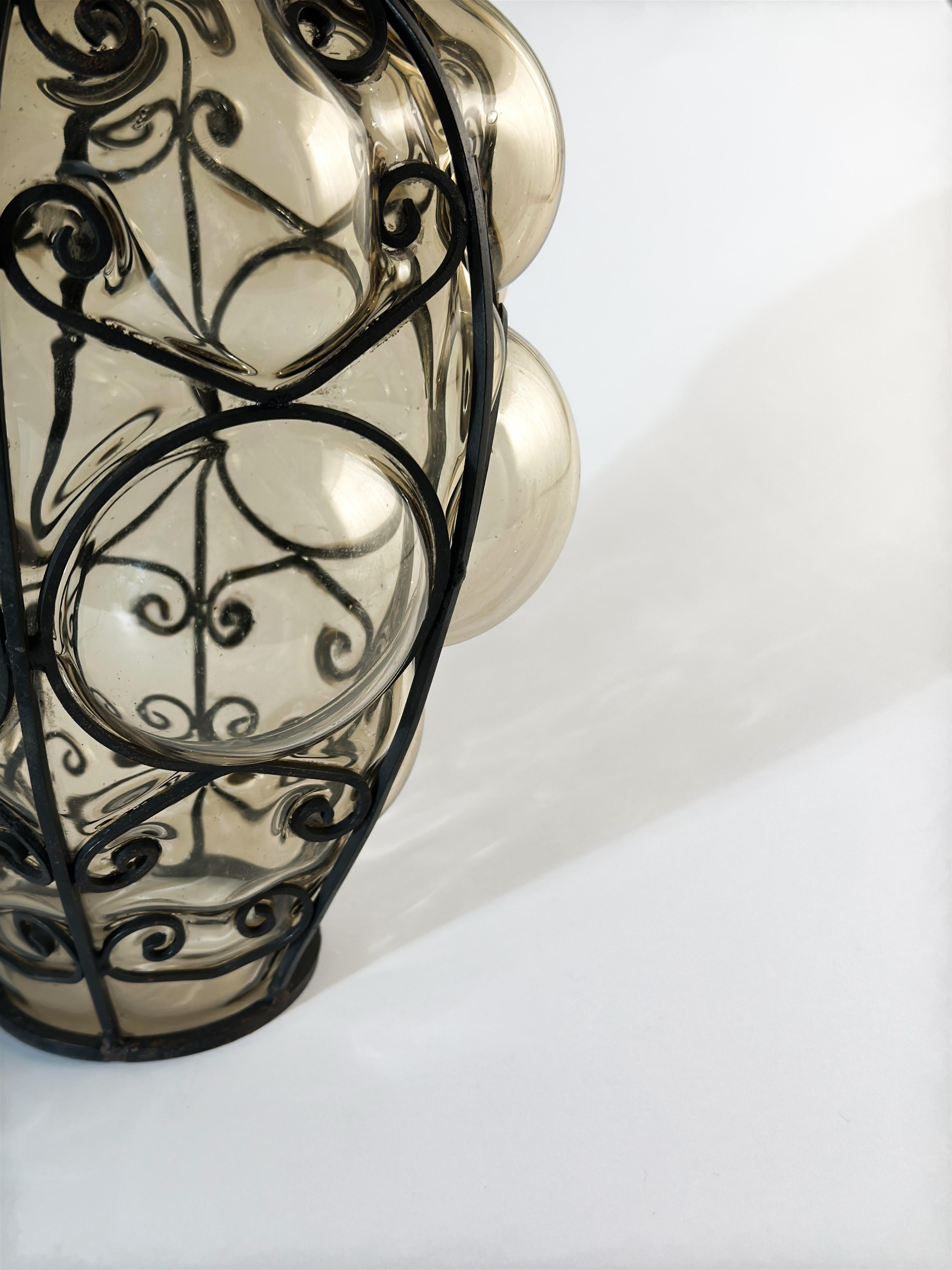 A Murano Glass caged table lamp circa 1950s, Italy

Exhibiting a compelling display of glassmaking technique, this caged glass lamp features a hand-forged wrought iron frame in a blackened finish. The rigid structure rests atop a layer of smoked