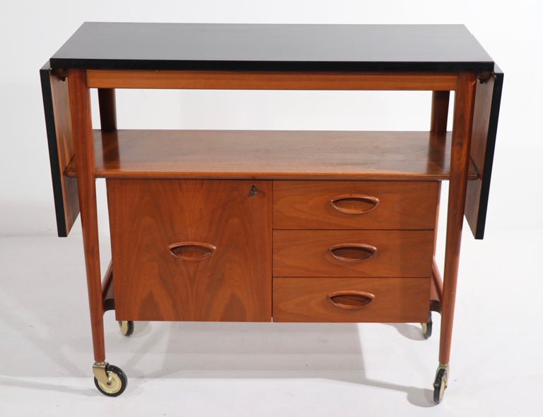 Exceptional Mid Century serving, bar cart with drop leaf extensions. The top is of black laminate, closed w 35.5 x open w 62 inch. The cart is of solid wood, with three drawers which flank a door that opens to additional storage. This example is in