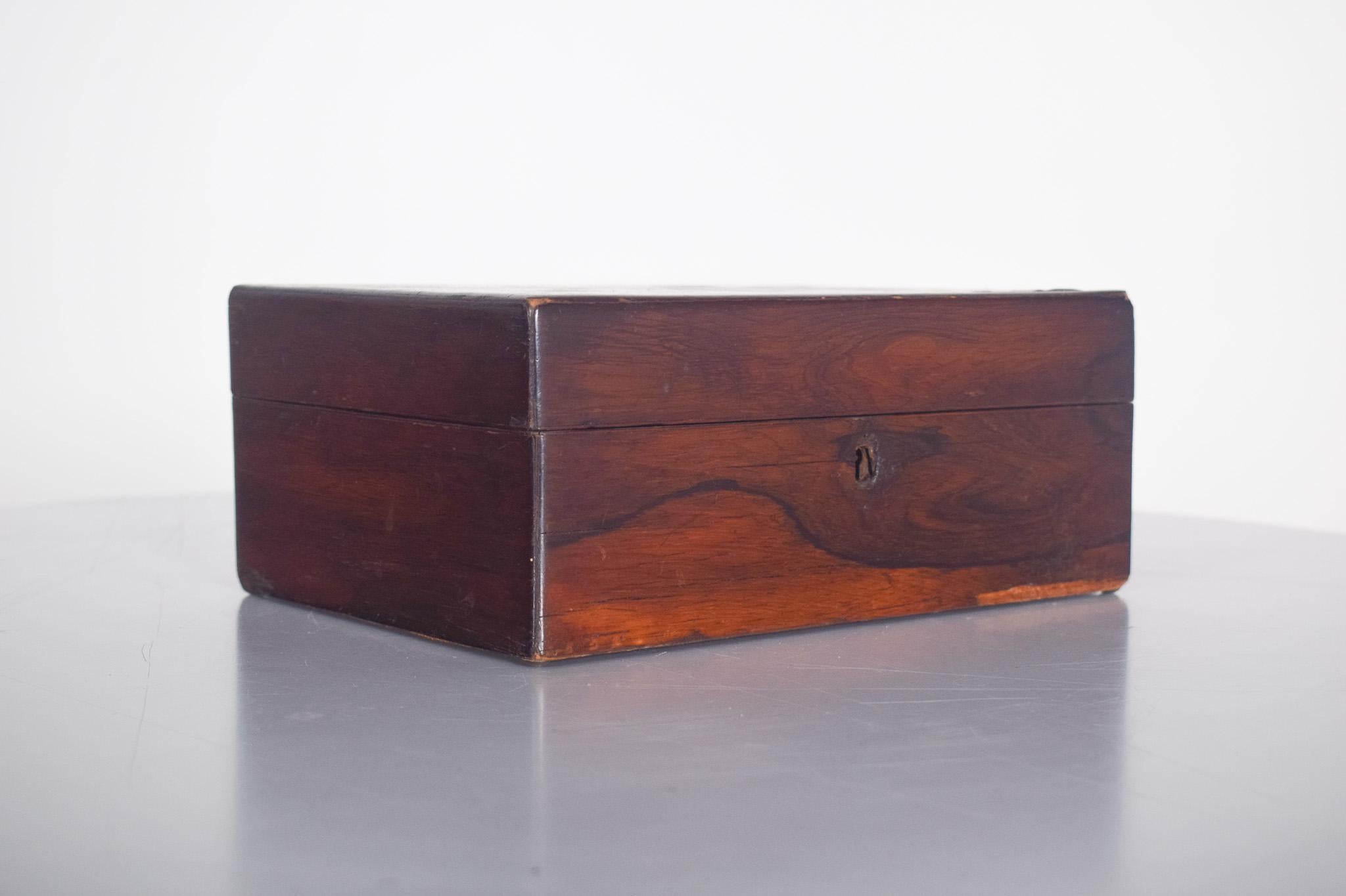 Costurero de palisandro. De madera oscura y con un acabado refinado, resalta las vetas naturales y da un toque elegante y clásico a la pieza. La estructura rectangular es robusta y está bien elaborada, con un pequeño cierre metálico en la parte