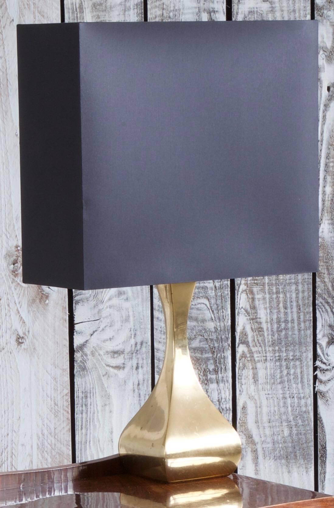 Midcentury table light with an elegantly curved solid brass base which contrasts with a strong black, block shaped lampshade which has a slim side depth to further emphasize the overall play in proportions. The inside of the shade has a gold foil