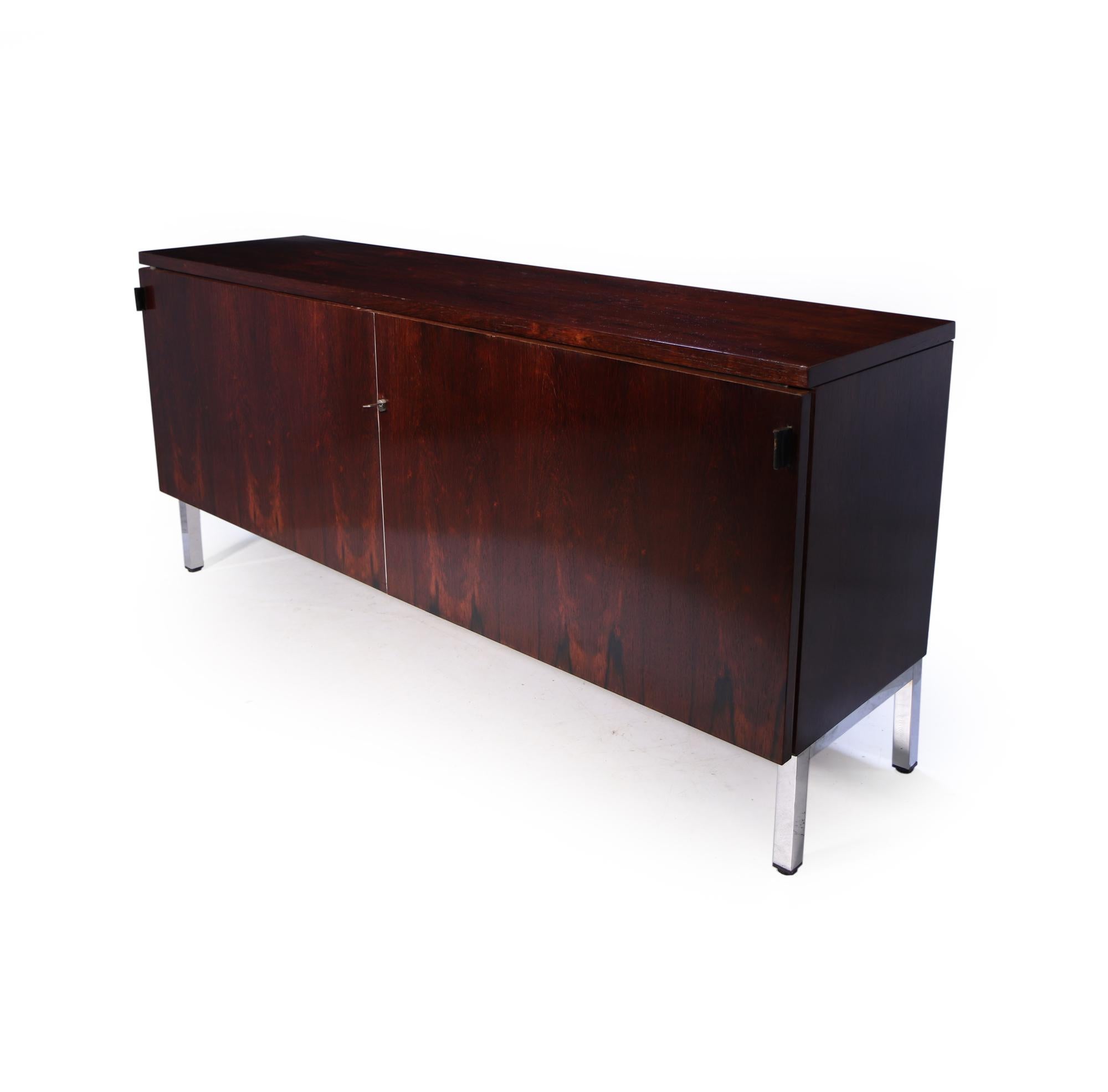 one of the most sought after iconic designs from the mid century modern era is this minimalist rosewood sideboard with two sliding doors with leather handles, and standing on a chrome base, two adjustable shelves, the sideboard has been fully